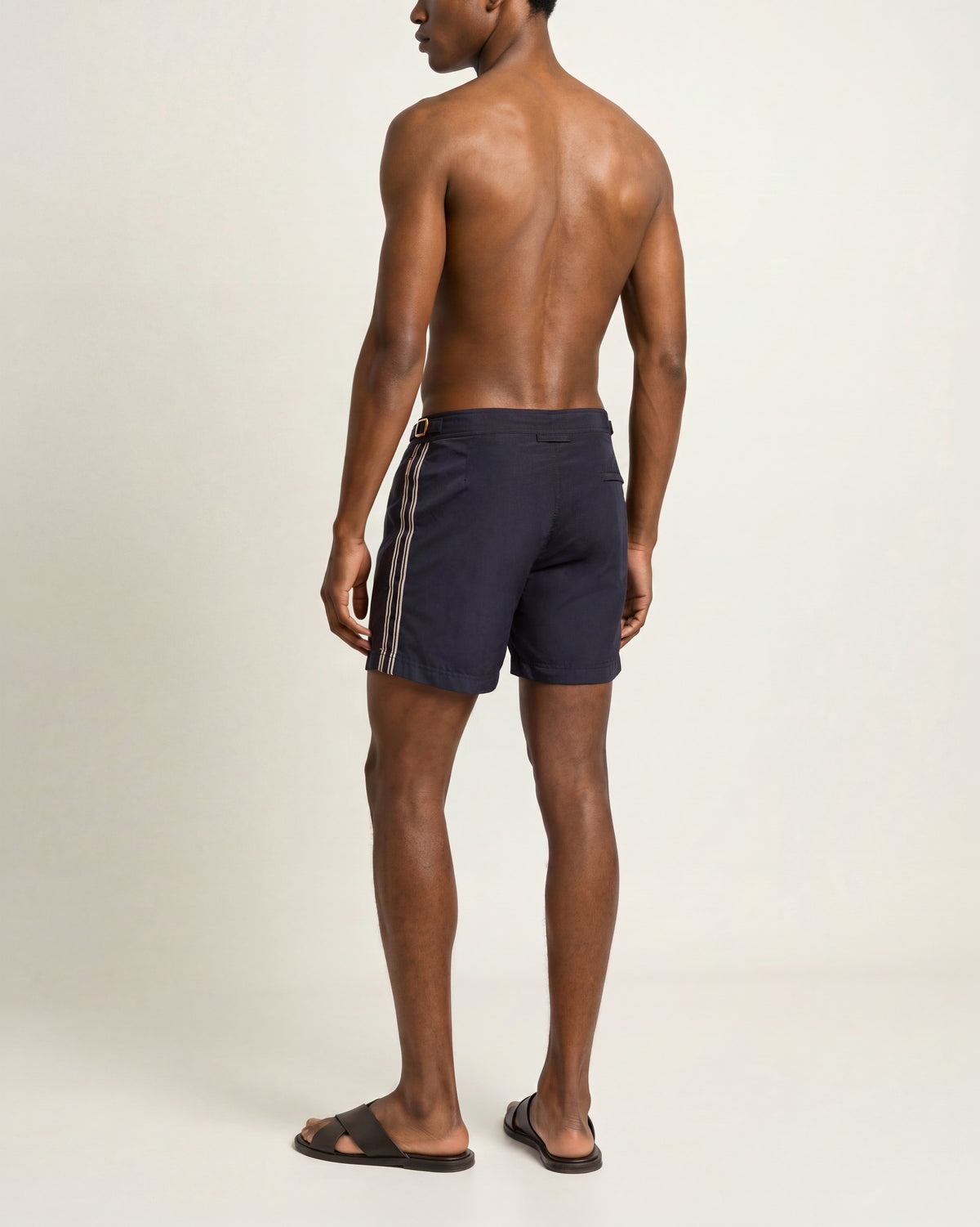 Back view of a man wearing navy swim shorts with a white stripe on a plain background