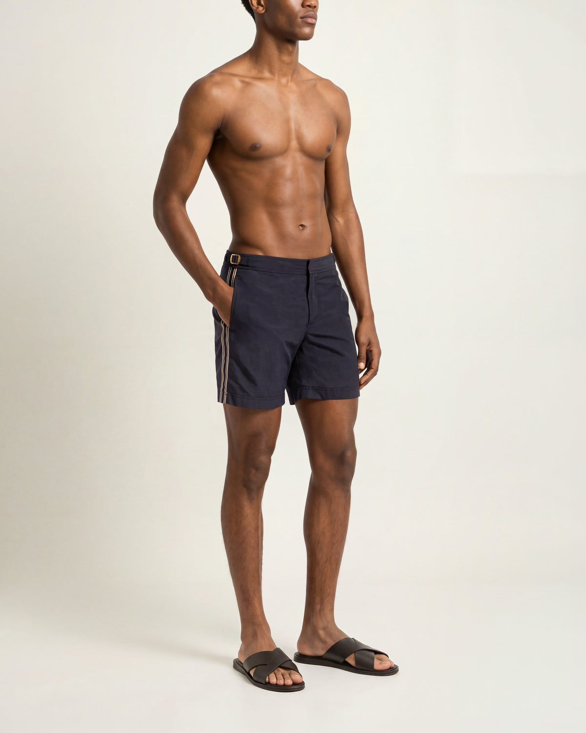 Man wearing navy swim shorts with a white stripe and brown sandals on a plain background