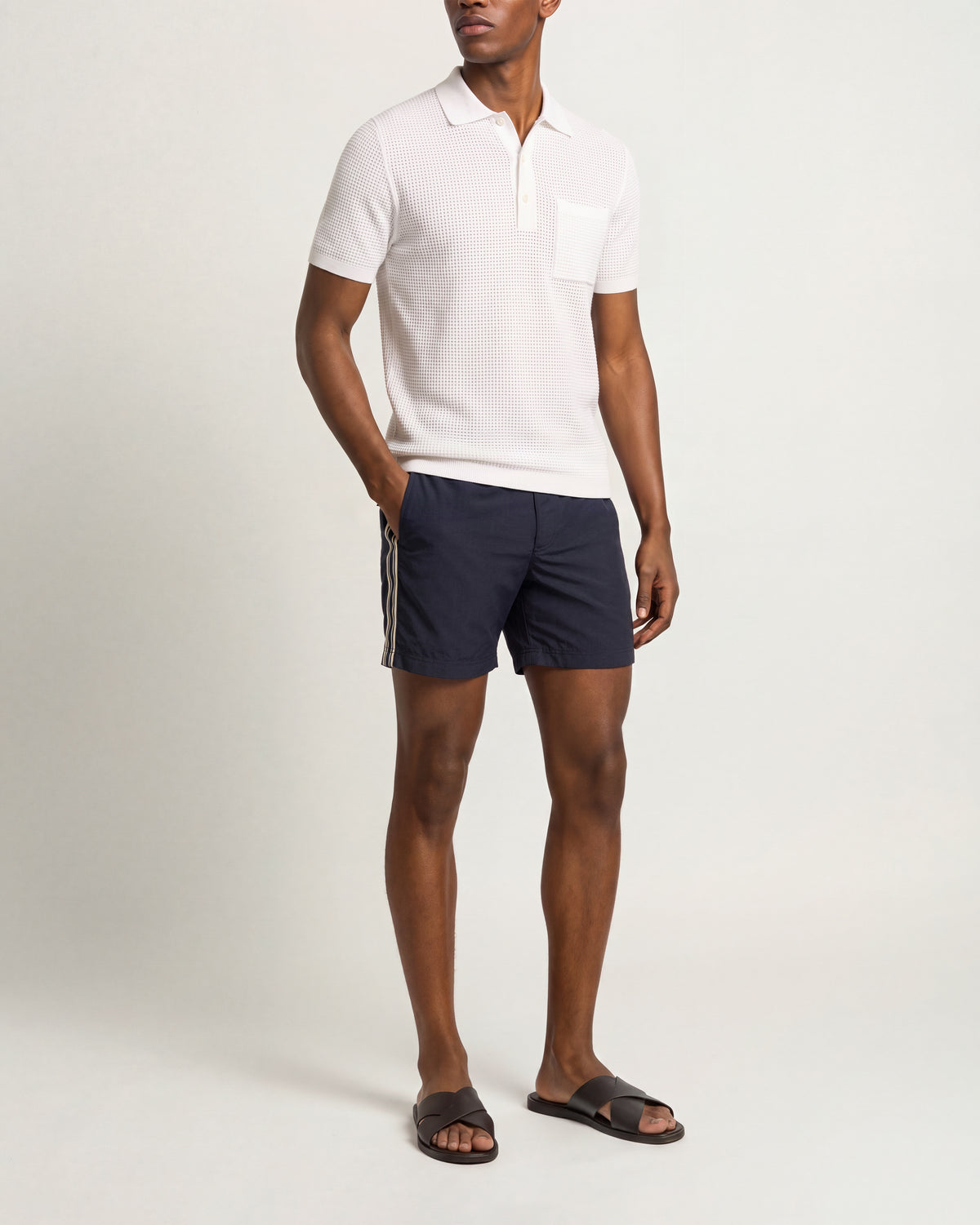 A man modeling a white short-sleeve polo shirt with a chest pocket, paired with navy blue shorts featuring white side stripes, and black slide sandals, against a plain light gray studio background.