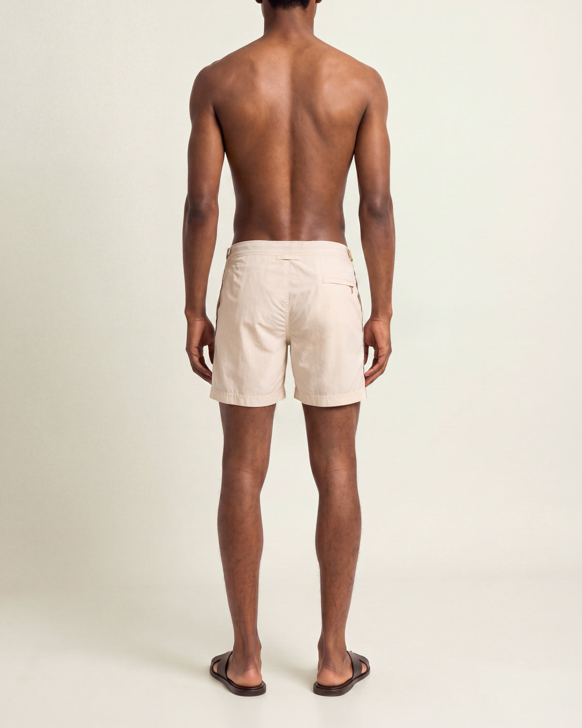 Man viewed from the back wearing a beige shorts with black stripes with brown sandals on a plain background
