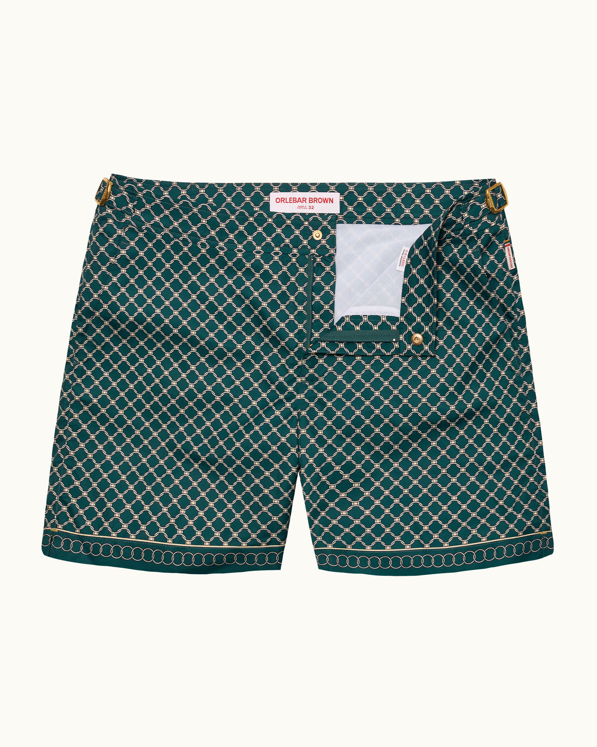 A pair of dark green patterned swimshorts with a white geometric lattice design, laid flat against a light cream background. The shorts feature a white inner lining visible in the pocket and a small red brand label at the waistband.