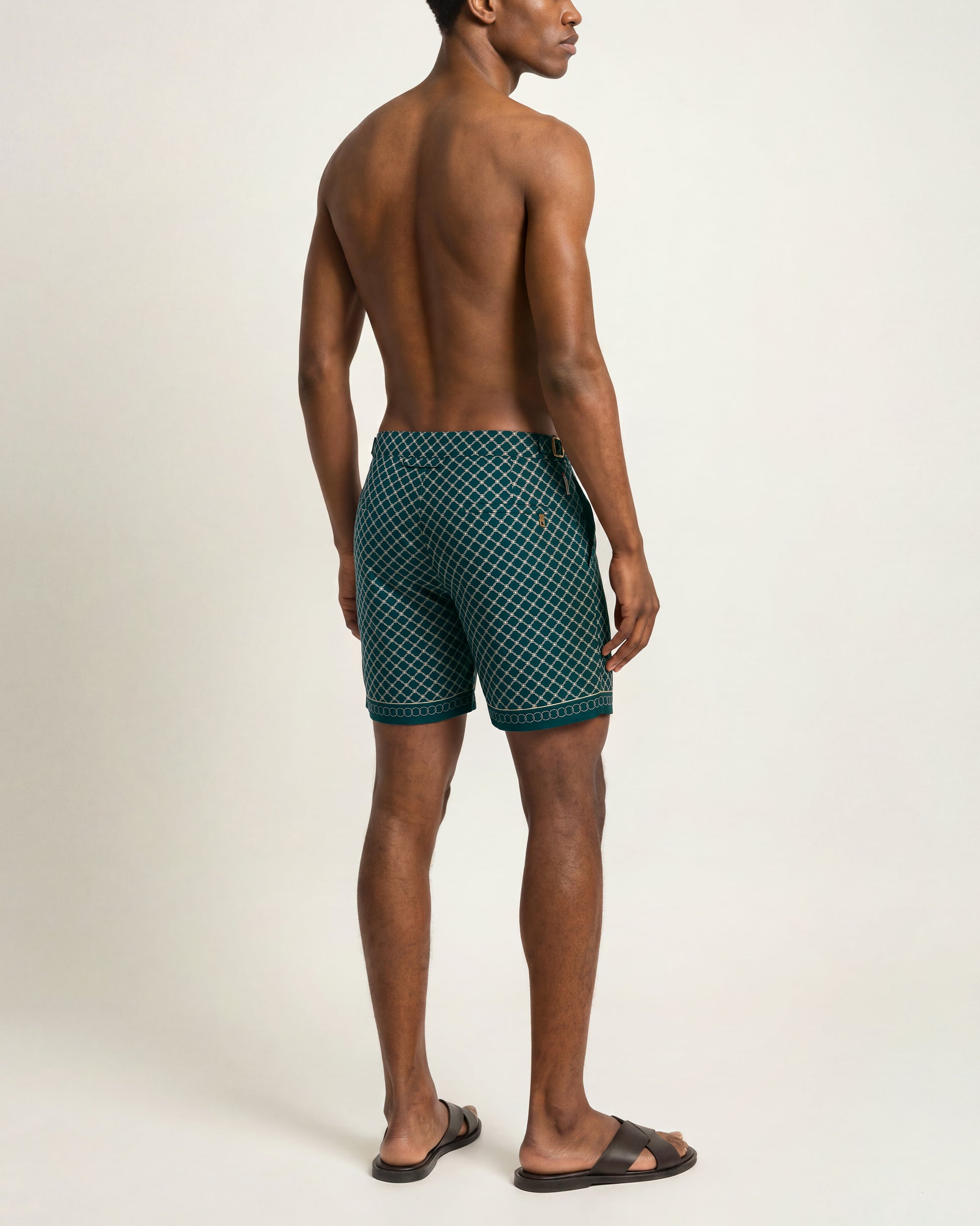 A man shown from the back wearing dark green patterned swim trunks and black slide sandals against a plain light beige studio background.