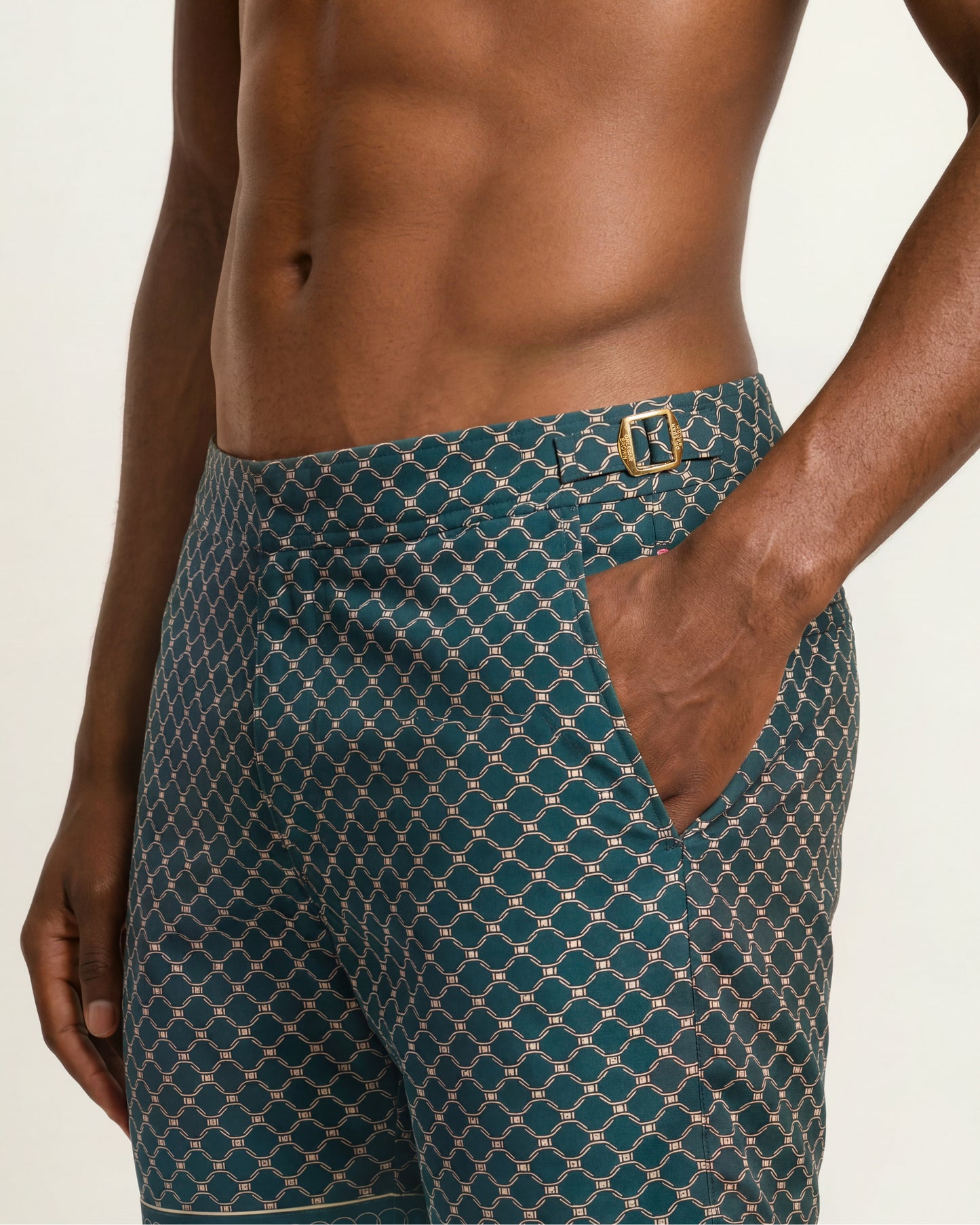 A close-up of a man wearing teal patterned swim trunks with a gold buckle detail, set against a plain light background.