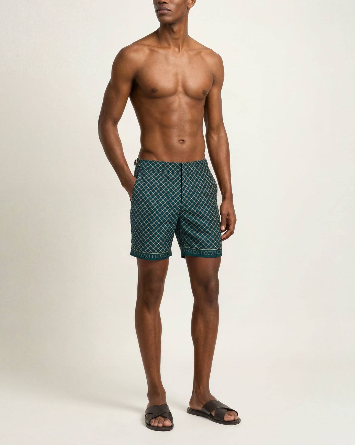 A shirtless male model wearing dark teal patterned swim trunks and dark brown slide sandals, standing against a plain light beige studio background.