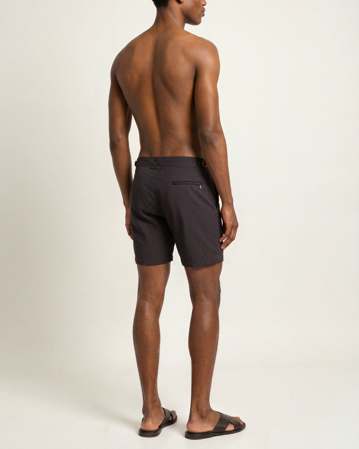 A man shown from the back wearing dark charcoal gray swim trunks and black slide sandals, standing against a plain off-white studio background.