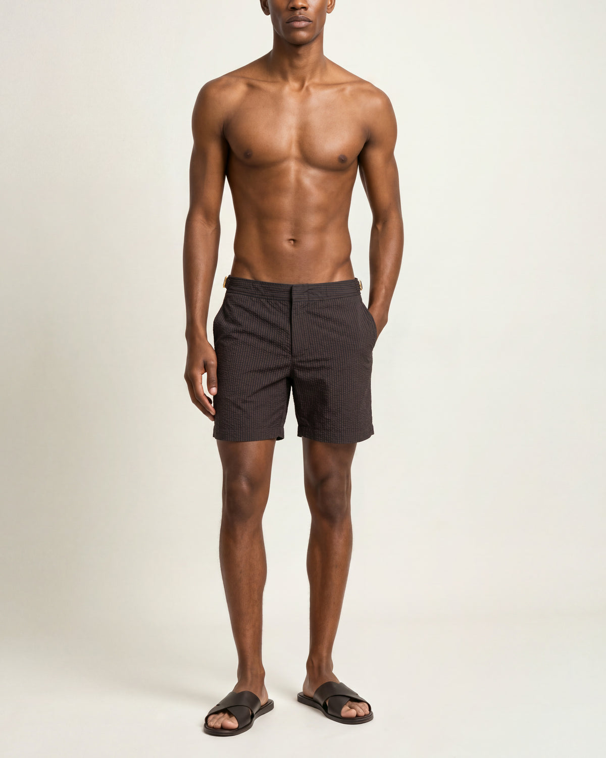A shirtless male model wearing dark brown shorts and black slide sandals, standing against a plain off-white studio background.