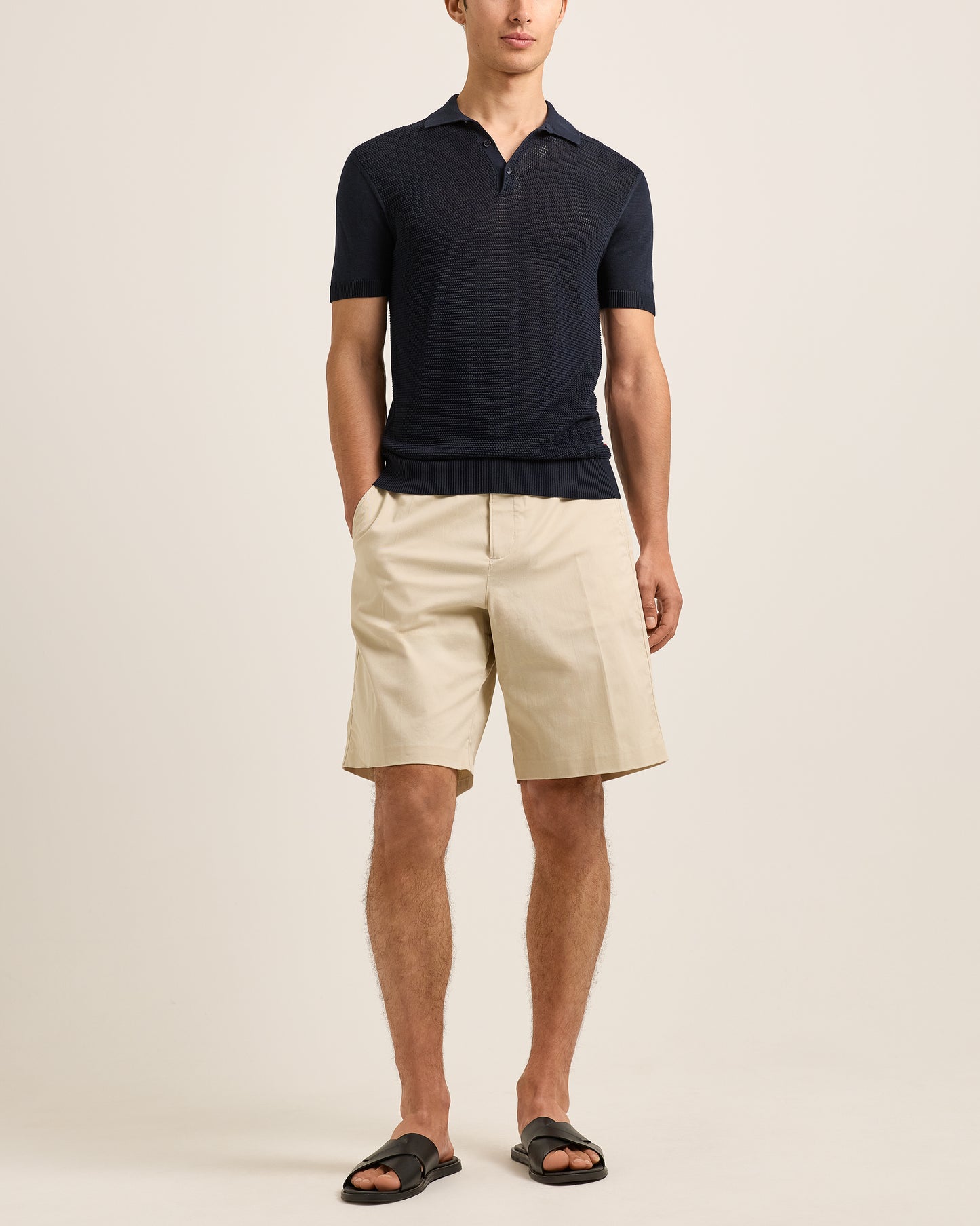 Man wearing a navy polo shirt and beige shorts on a plain background