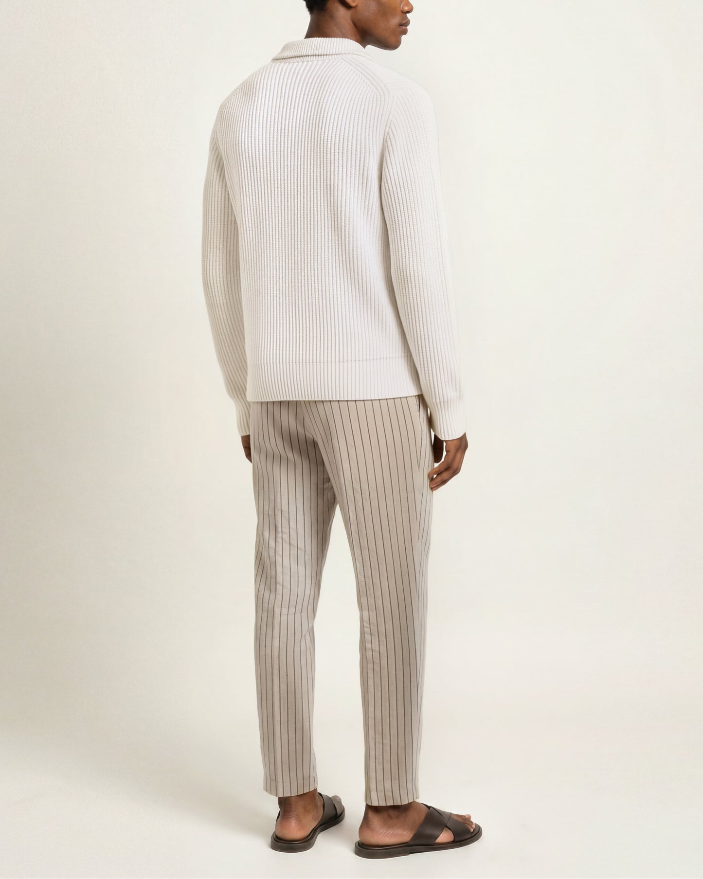A man shown from the back wearing a cream ribbed knit polo sweater and beige pinstriped trousers, paired with dark brown sandals, against a plain off-white studio background.