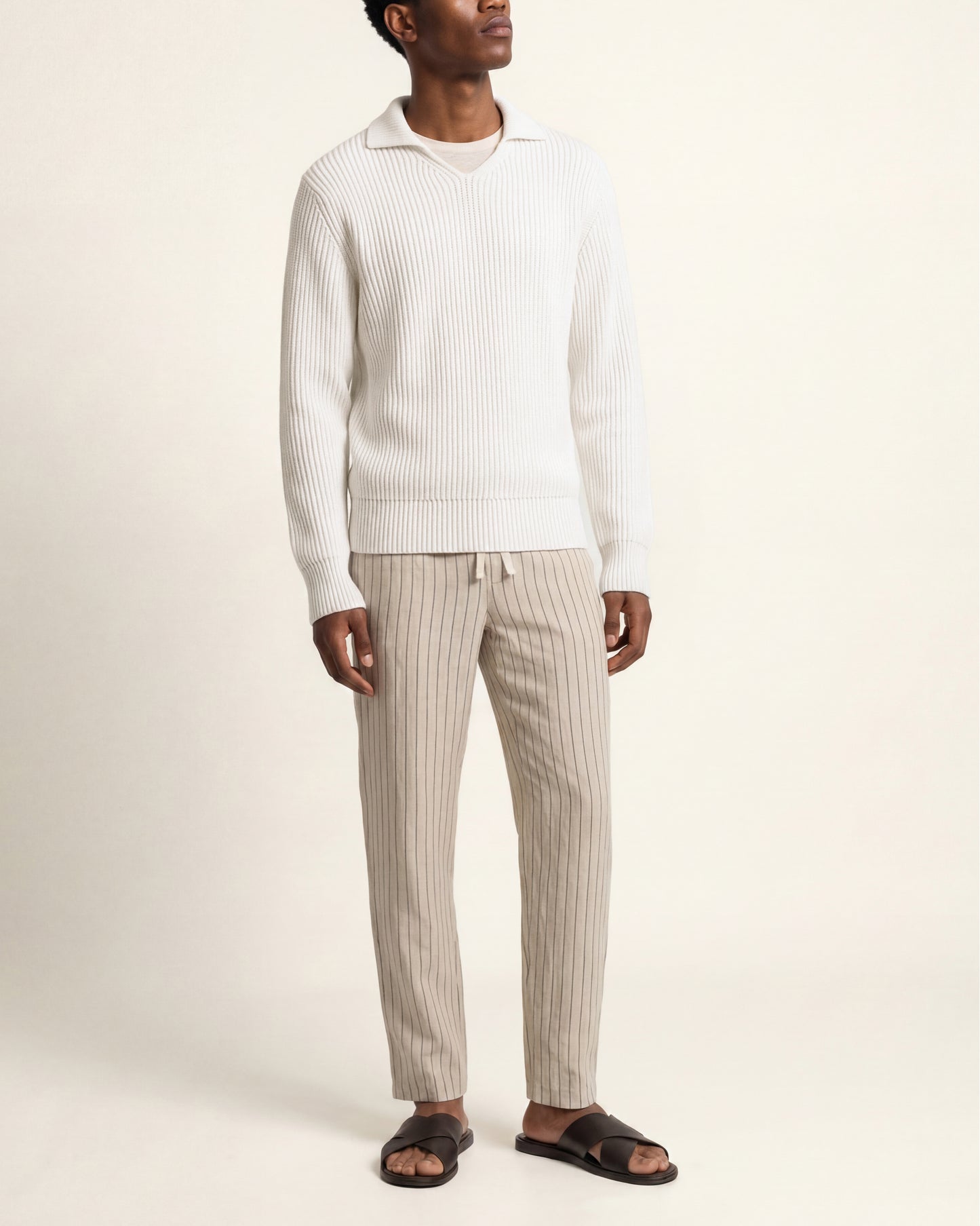 A man wearing a white ribbed knit sweater and beige vertically striped knit pants with dark brown slide sandals, standing against a plain cream background.