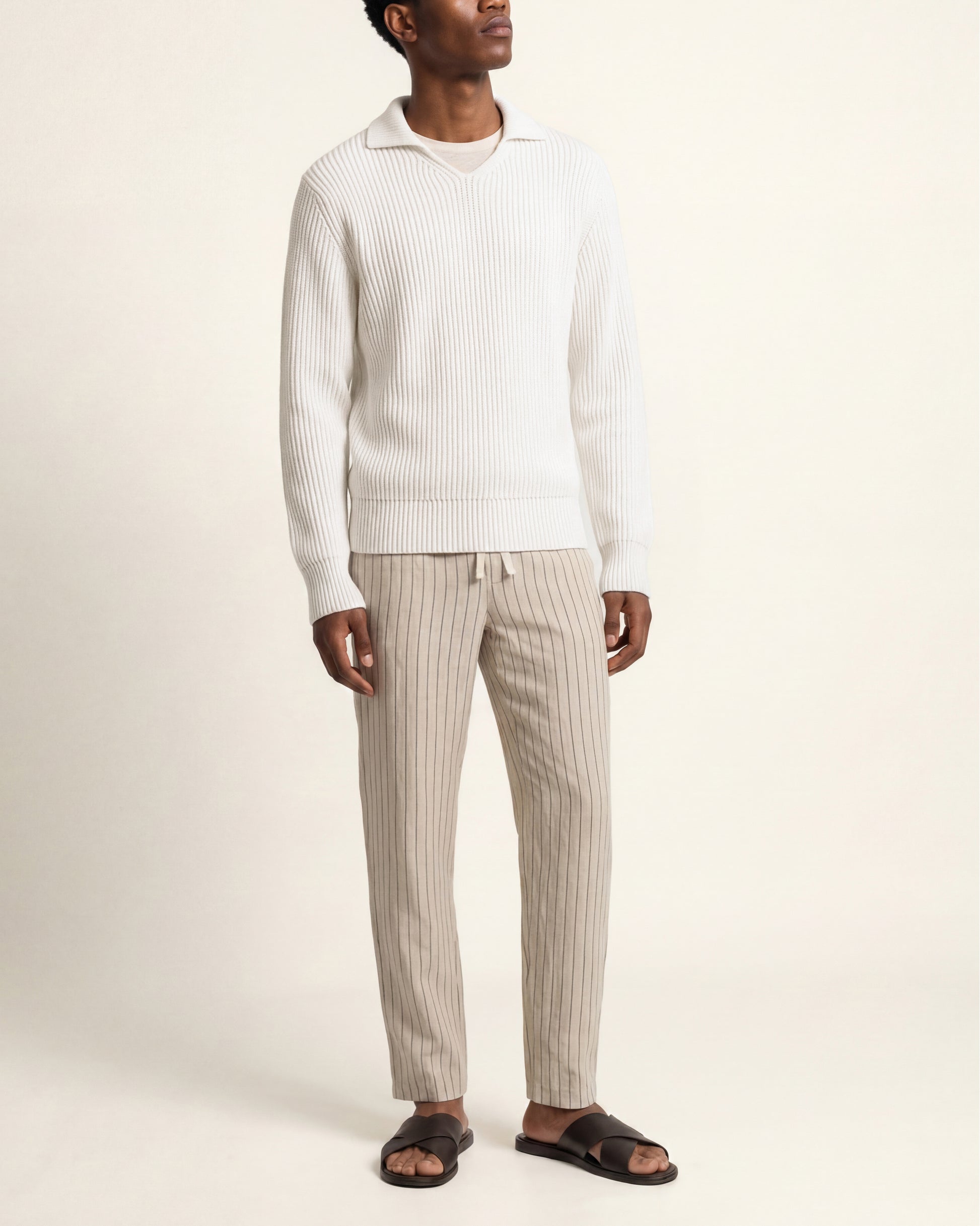 A man wearing a white ribbed knit sweater and beige vertically striped knit pants with dark brown slide sandals, standing against a plain cream background.