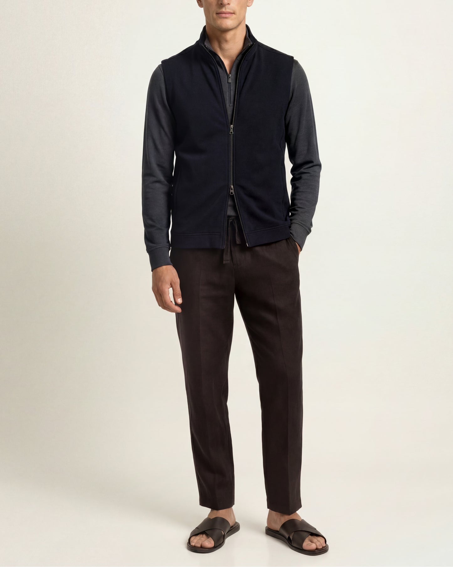 A man modeling a dark navy sleeveless zip-front vest over a dark long-sleeve shirt, paired with dark brown trousers and black sandals, against a plain light background.