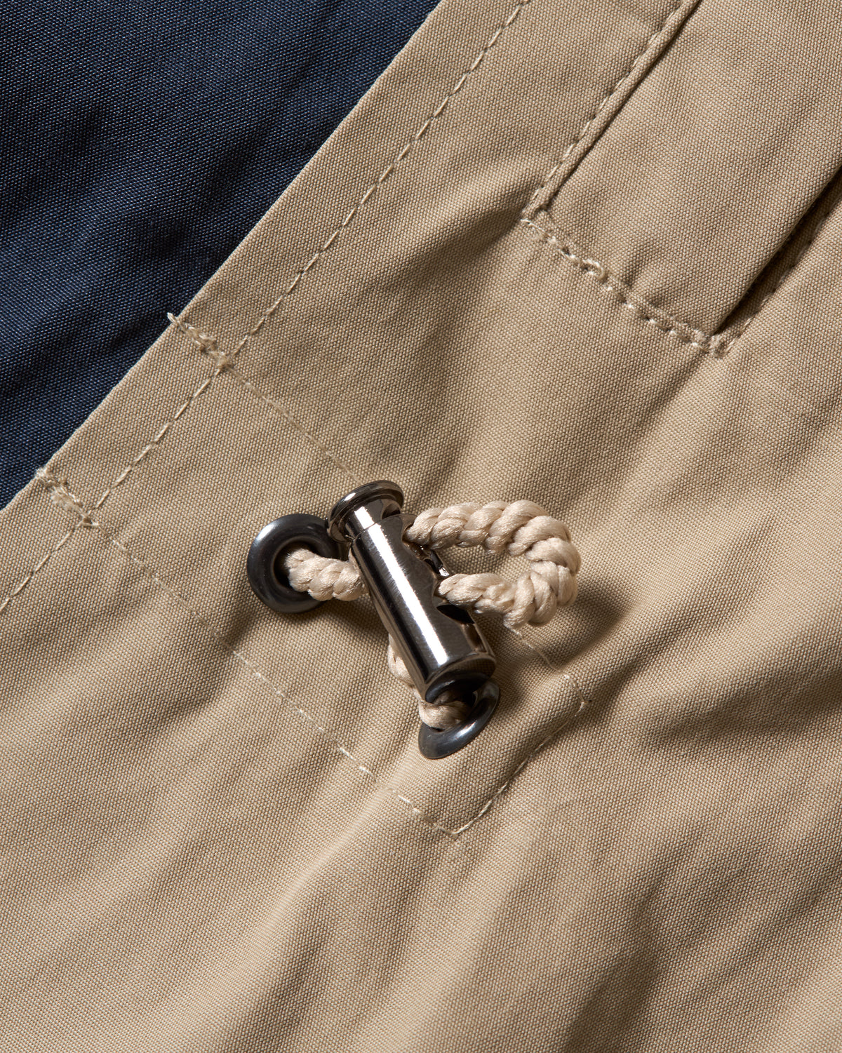 Close-up of a pair of beige jacket showing the waistband, a side pocket, and a silver metal toggle with a beige rope closure.