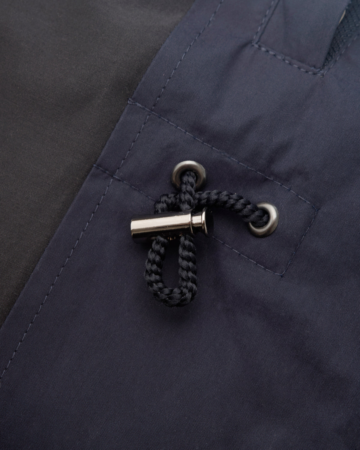 Close-up of a dark navy jacket or waterproof outerwear detail, showing a black braided drawcord with a silver metal toggle and silver eyelets.