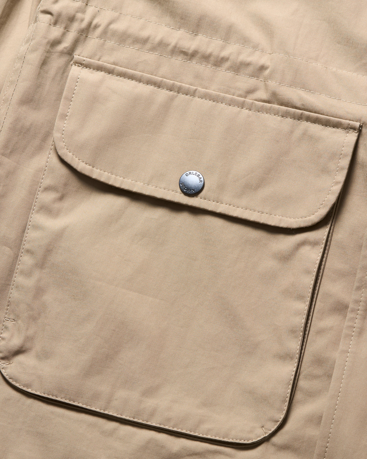 Close-up of a beige utility jacket or shirt featuring a large front flap pocket secured by a silver snap button.