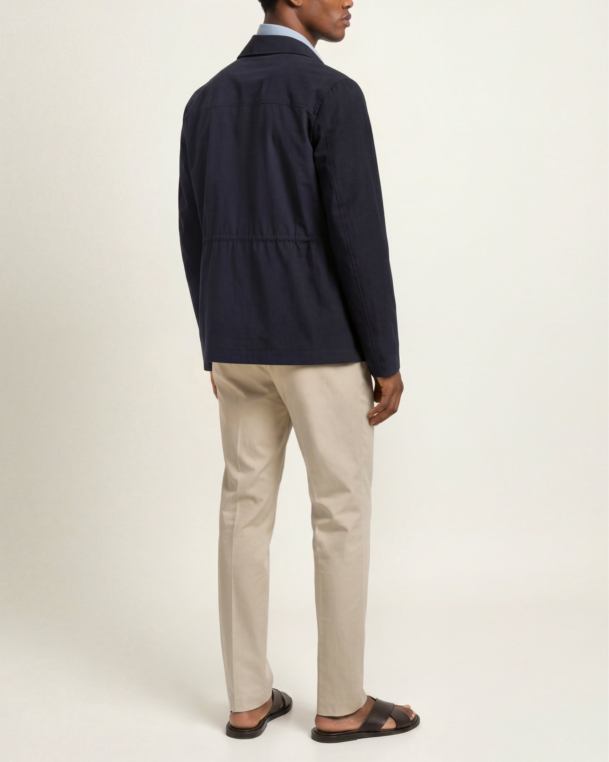A man shown from the back wearing a dark navy jacket, beige trousers, and dark brown sandals against a plain off-white studio background.