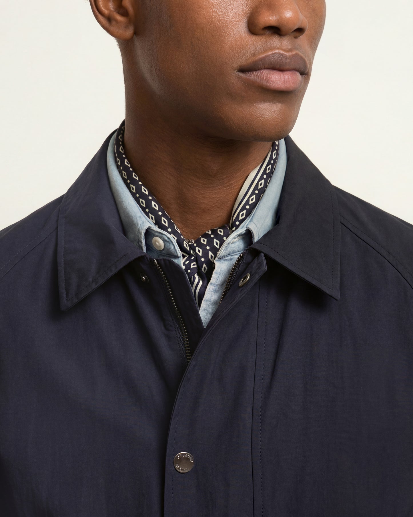 Close-up of a man wearing a dark navy jacket over a light blue collared shirt with a patterned neck scarf, against a plain light background.