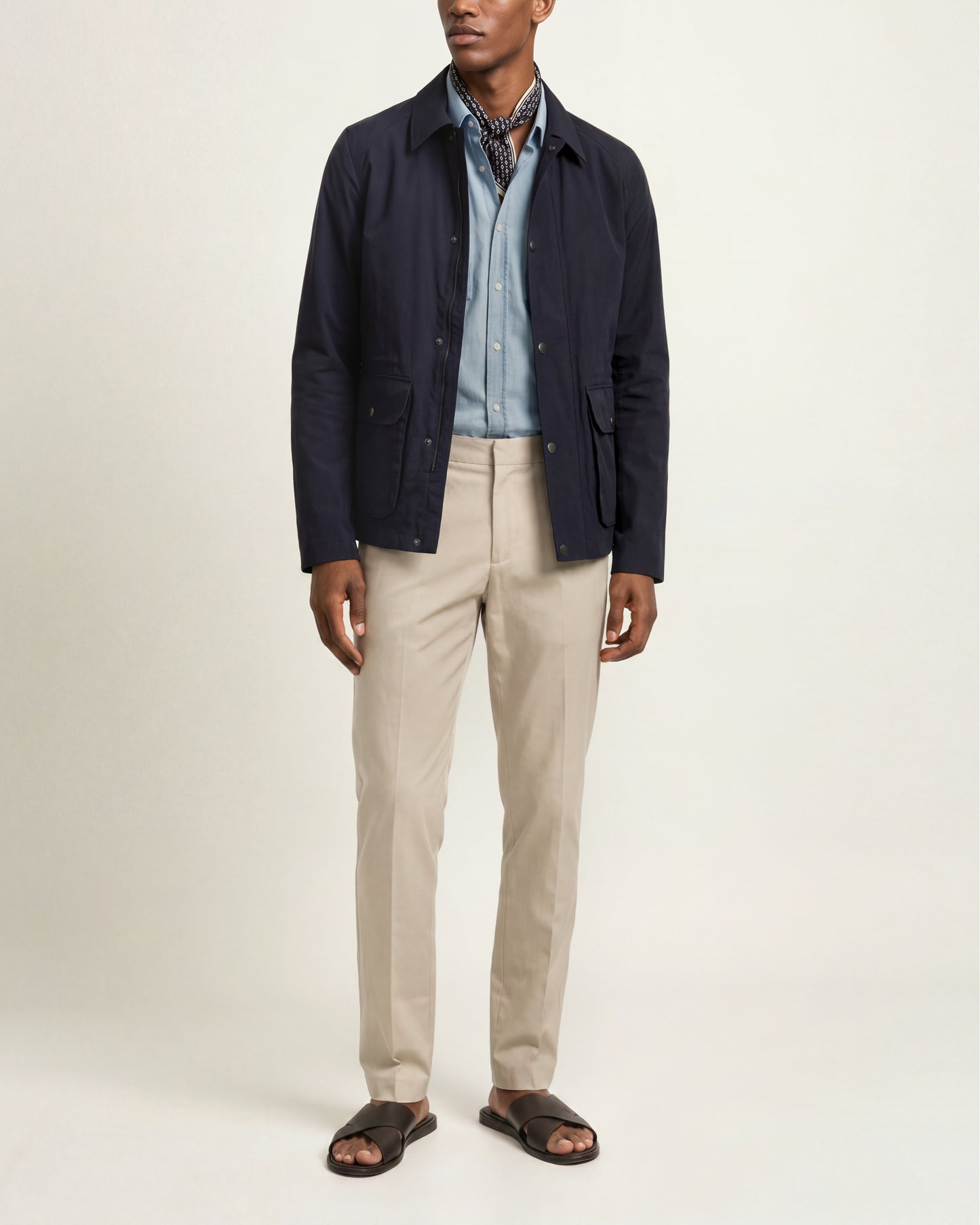 A man modeling a dark navy blue lightweight jacket over a light blue button-down shirt, paired with beige trousers and dark brown slide sandals, against a plain off-white studio background.
