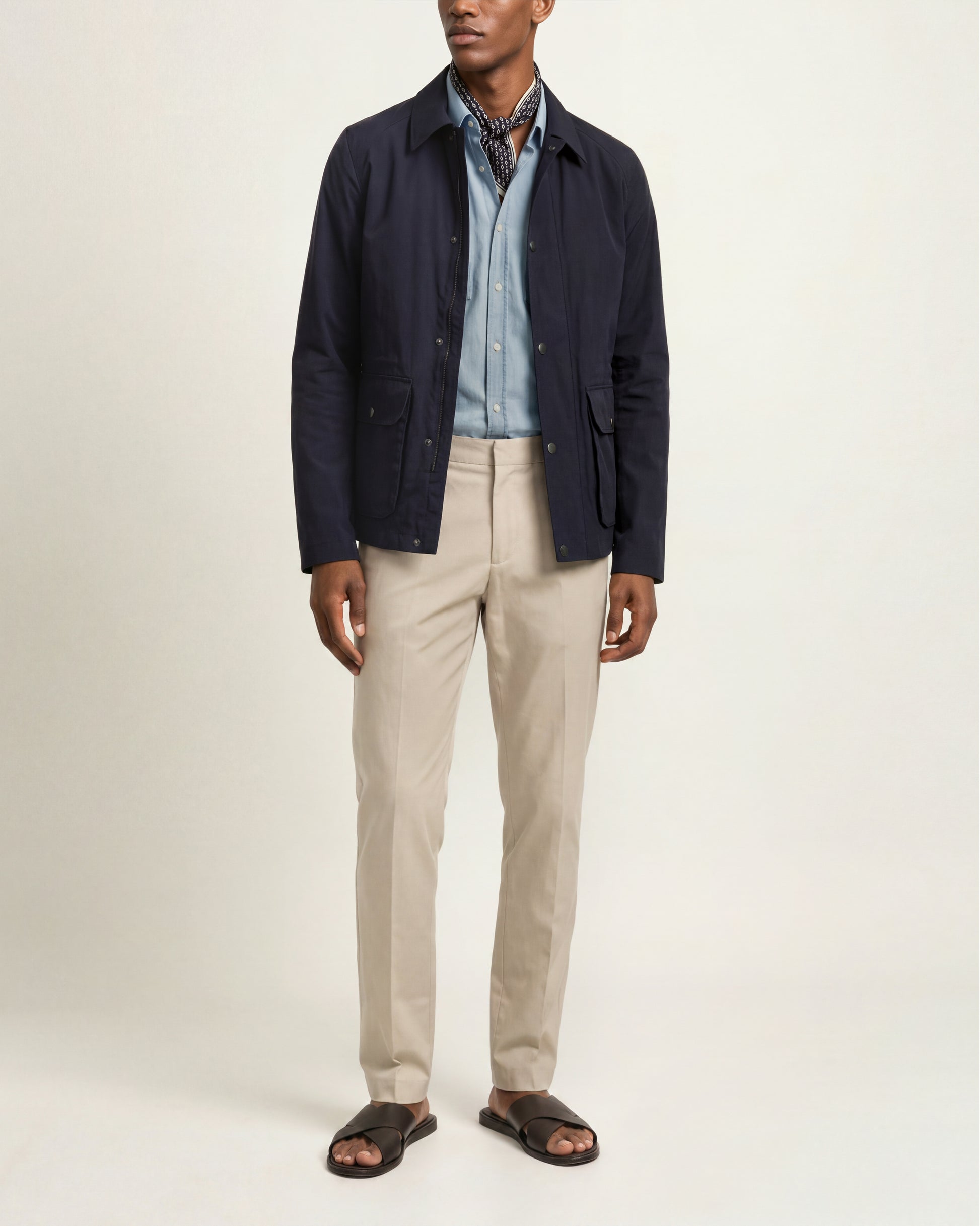 A man modeling a dark navy blue lightweight jacket over a light blue button-down shirt, paired with beige trousers and dark brown slide sandals, against a plain off-white studio background.