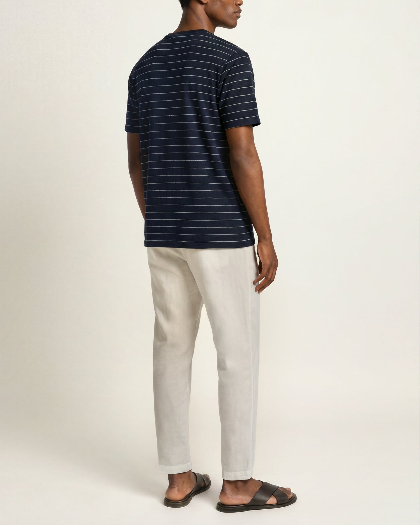 A man shown from the back wearing a navy blue short-sleeve t-shirt with thin white horizontal stripes, off-white trousers, and dark brown slide sandals, set against a plain light beige studio background.