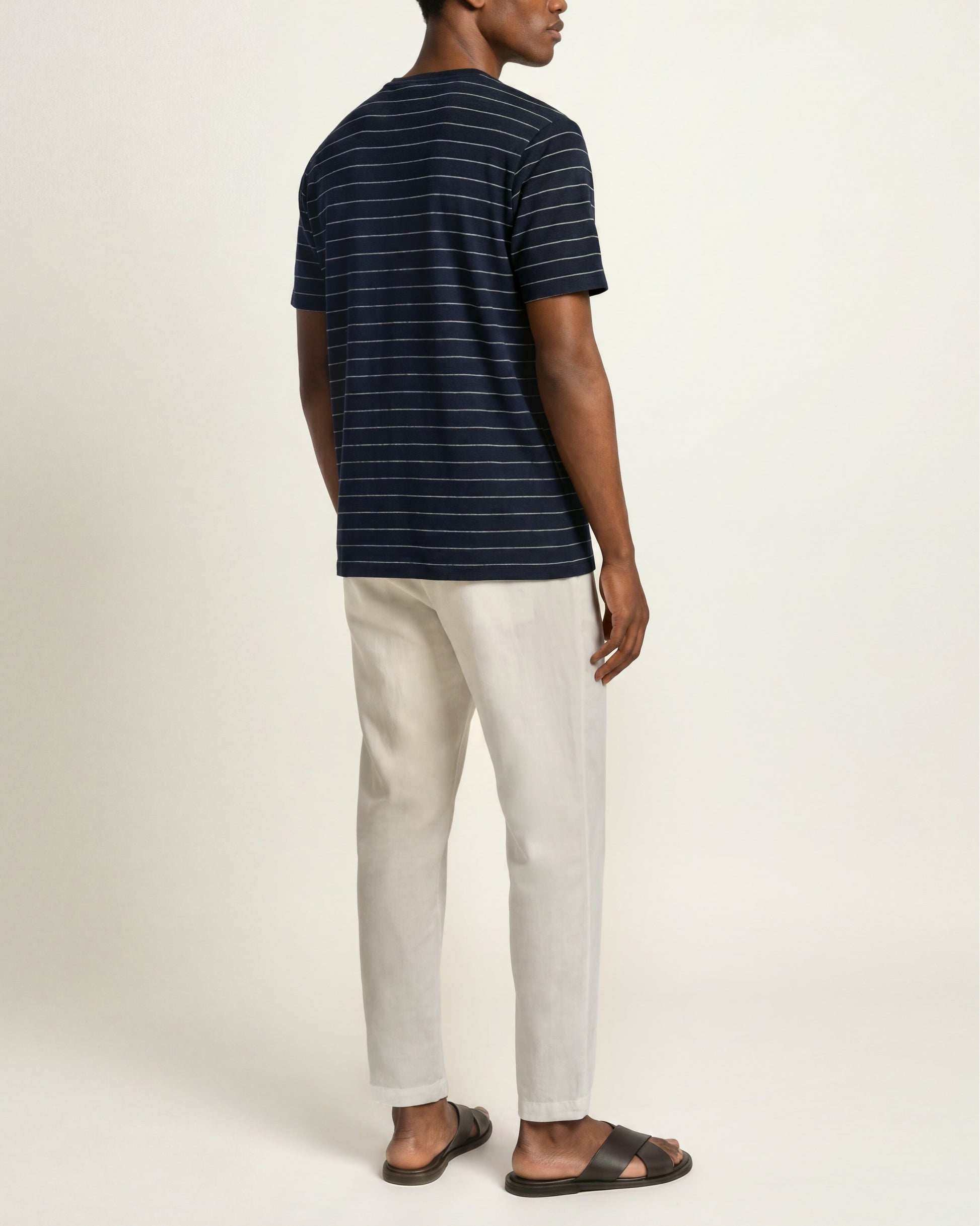 A man shown from the back wearing a navy blue short-sleeve t-shirt with thin white horizontal stripes, off-white trousers, and dark brown slide sandals, set against a plain light beige studio background.
