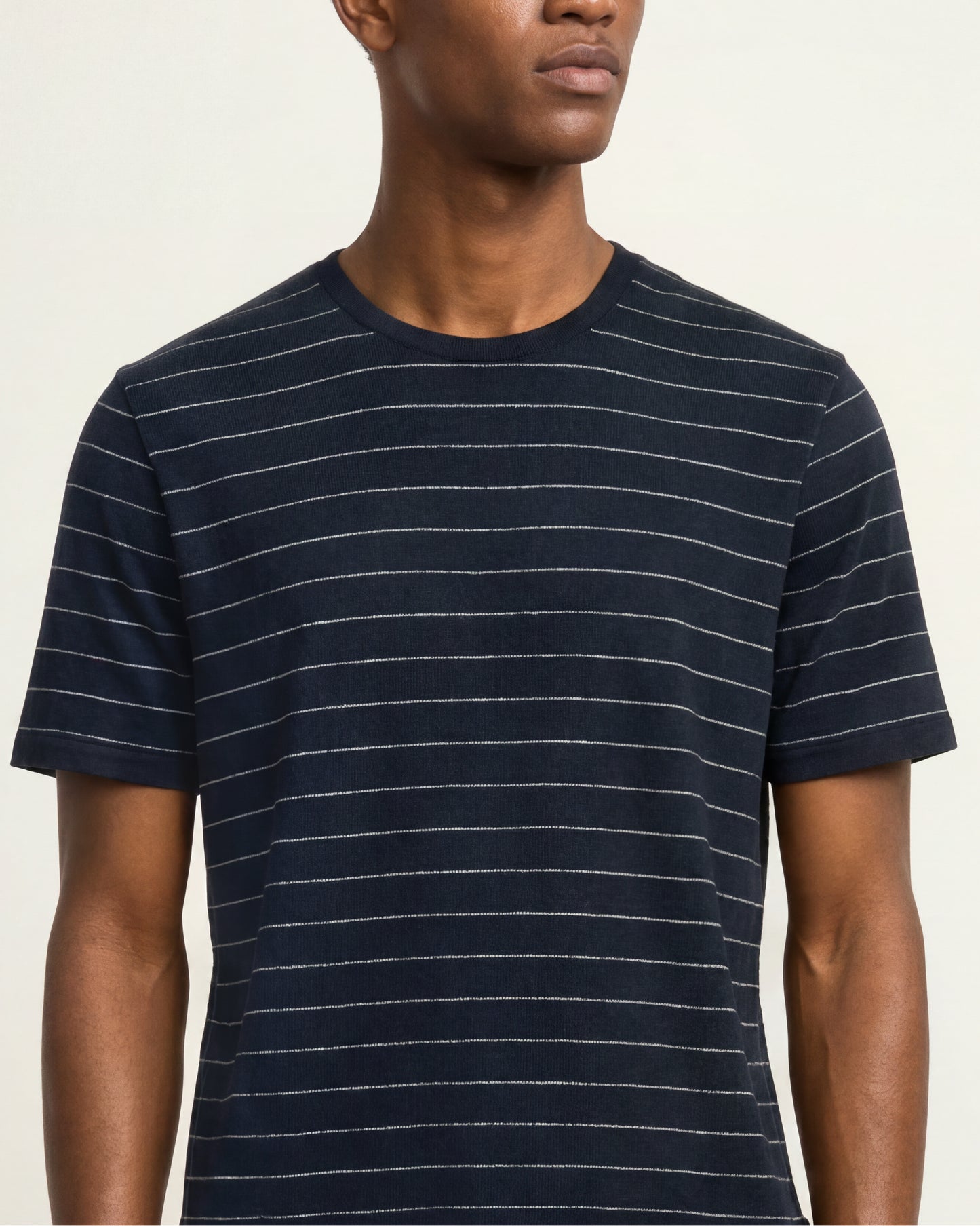 A man wearing a navy blue short-sleeve crewneck t-shirt with thin white horizontal stripes, shown from the chest up against a light beige background.