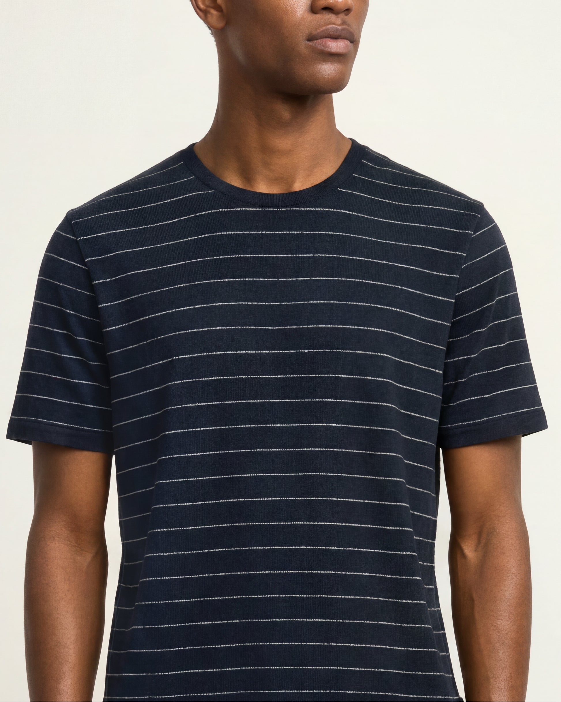 A man wearing a navy blue short-sleeve crewneck t-shirt with thin white horizontal stripes, shown from the chest up against a light beige background.