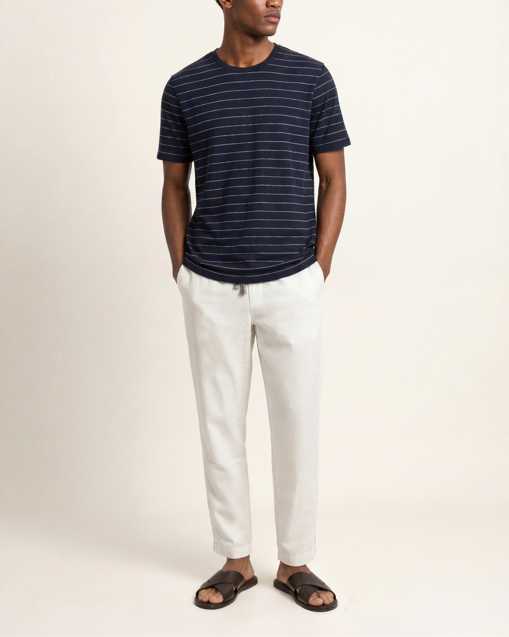 A man wearing a navy blue short-sleeve t-shirt with thin white horizontal stripes, paired with off-white trousers and dark brown slide sandals, standing against a plain light beige studio background.