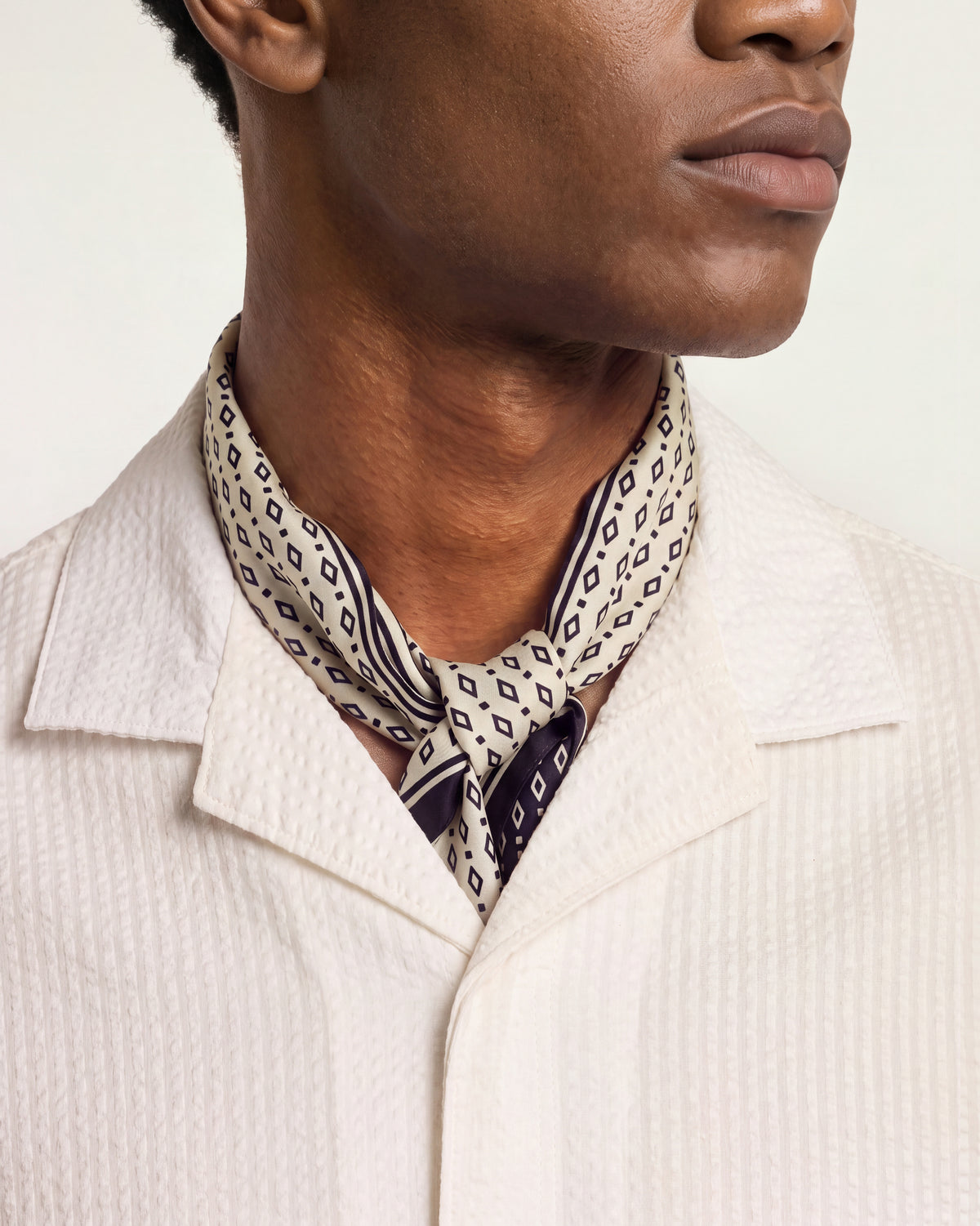 A close-up of a man wearing a cream-colored textured shirt and a patterned neck scarf with black and white geometric designs.