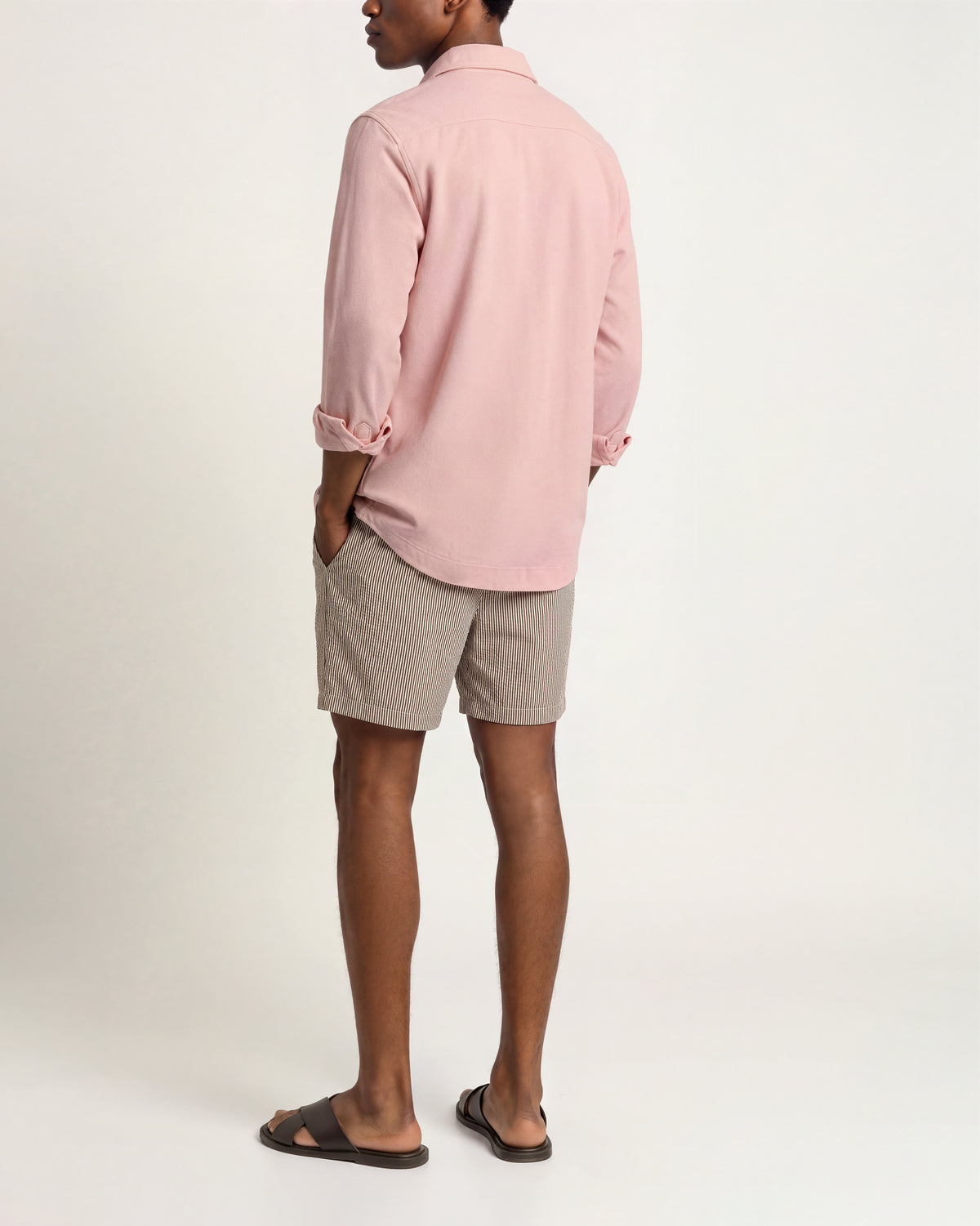 A man shown from the back wearing a light pink long-sleeve button-up shirt, light gray shorts, and dark brown slide sandals, standing against a plain off-white studio background.