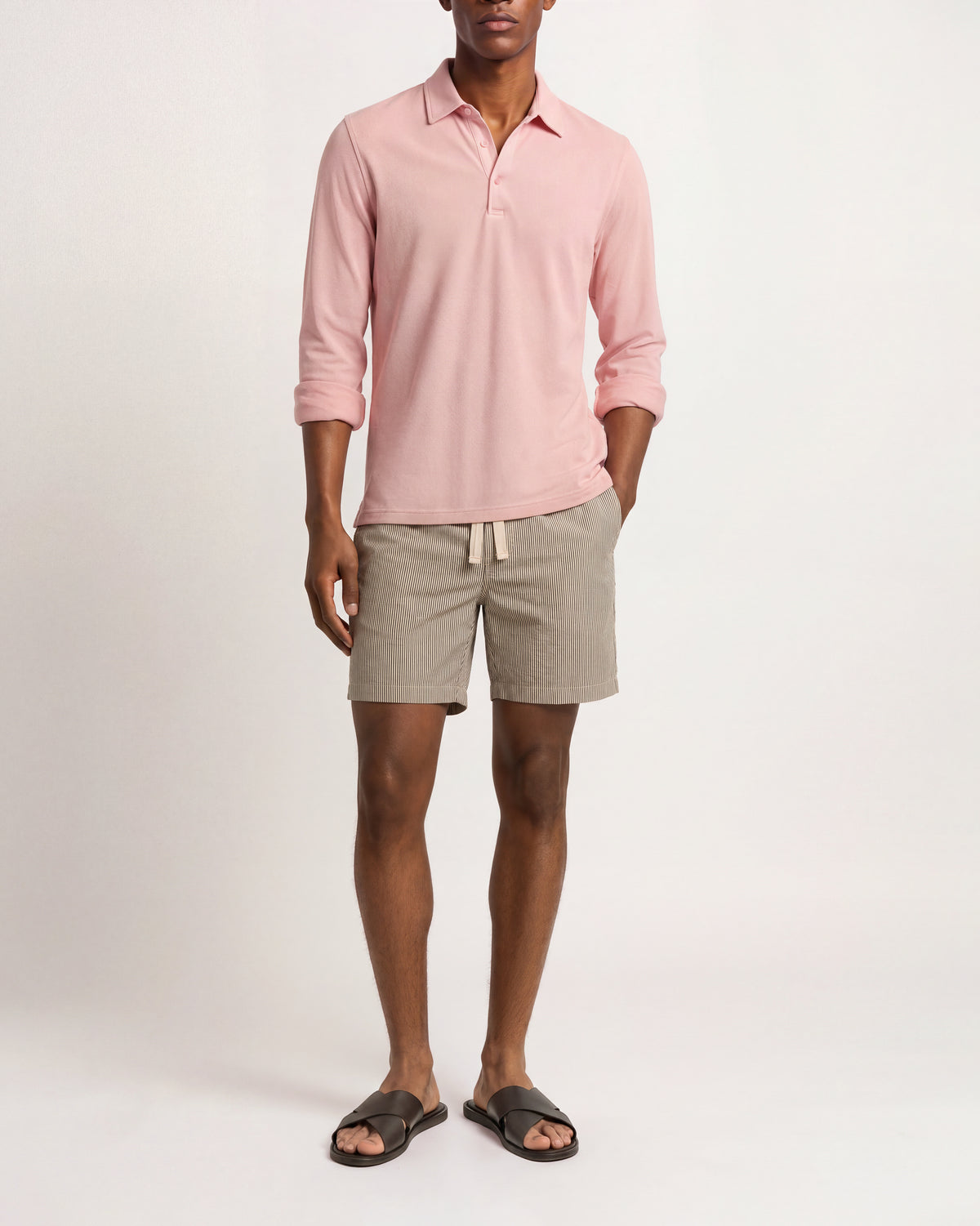 A man modeling a light pink long-sleeve polo shirt, light beige drawstring shorts, and dark brown slide sandals against a plain white studio background.