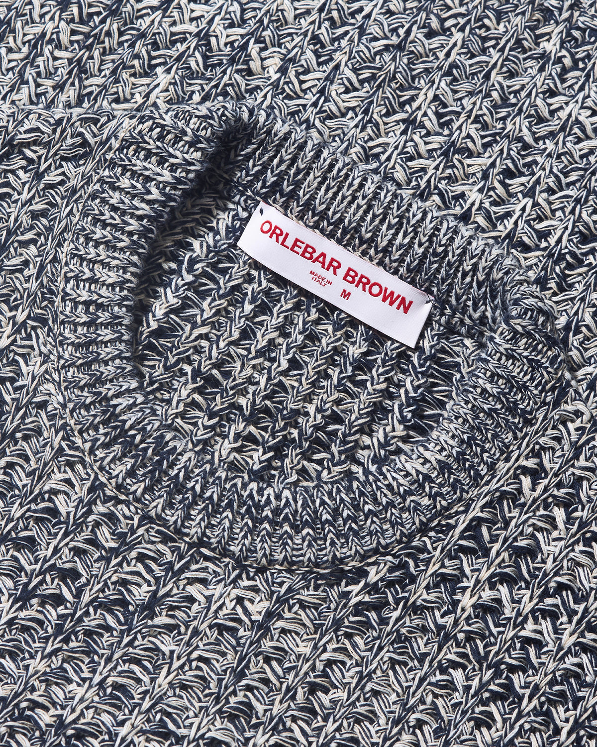 Close-up of a textured knit sweater with a white brand label reading ORLEBAR BROWN, showing a marled navy and off-white fabric.
