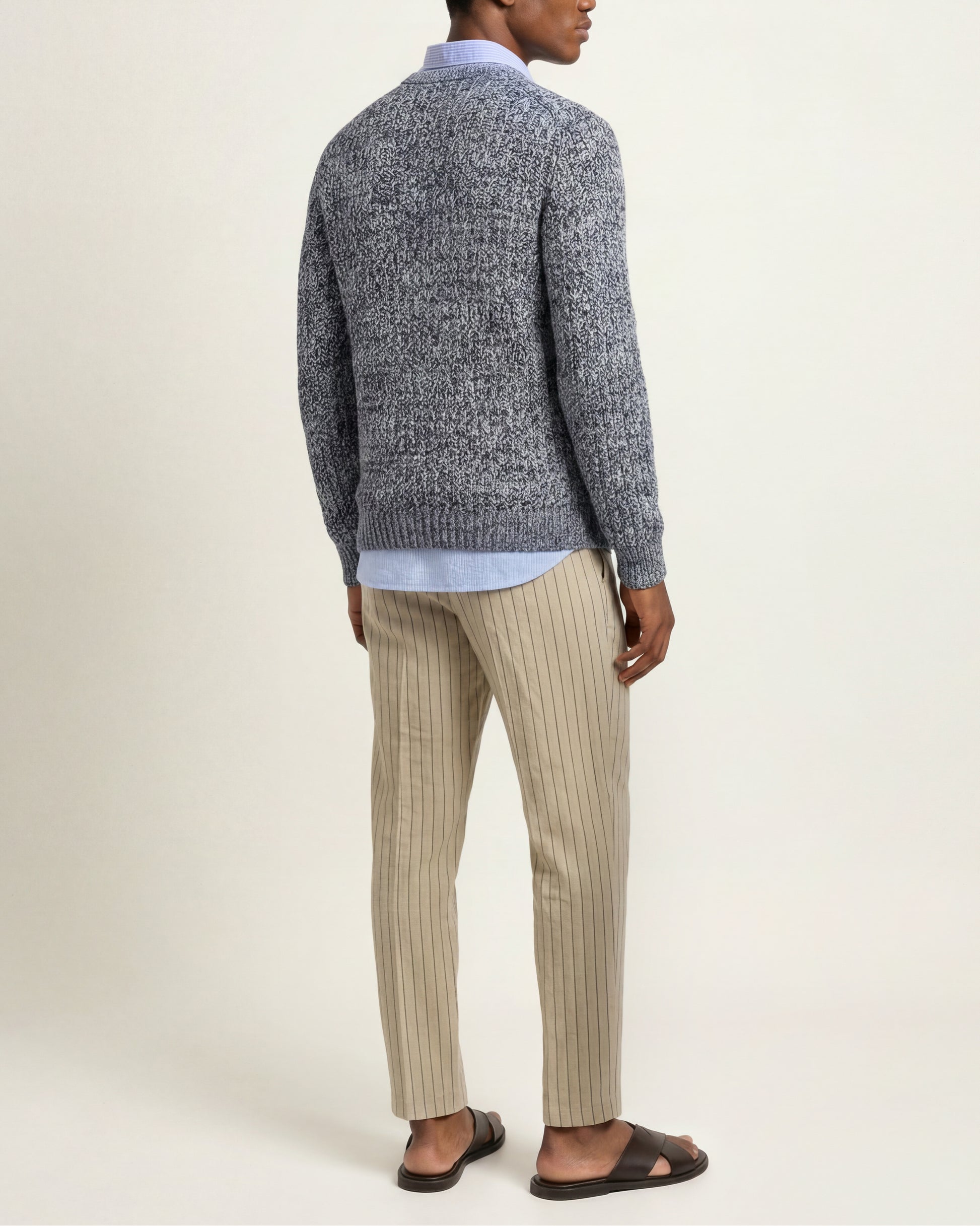A man shown from the back wearing a textured gray knit sweater, beige pinstriped trousers, a light blue collared shirt, and dark brown sandals, against a plain off-white studio background.