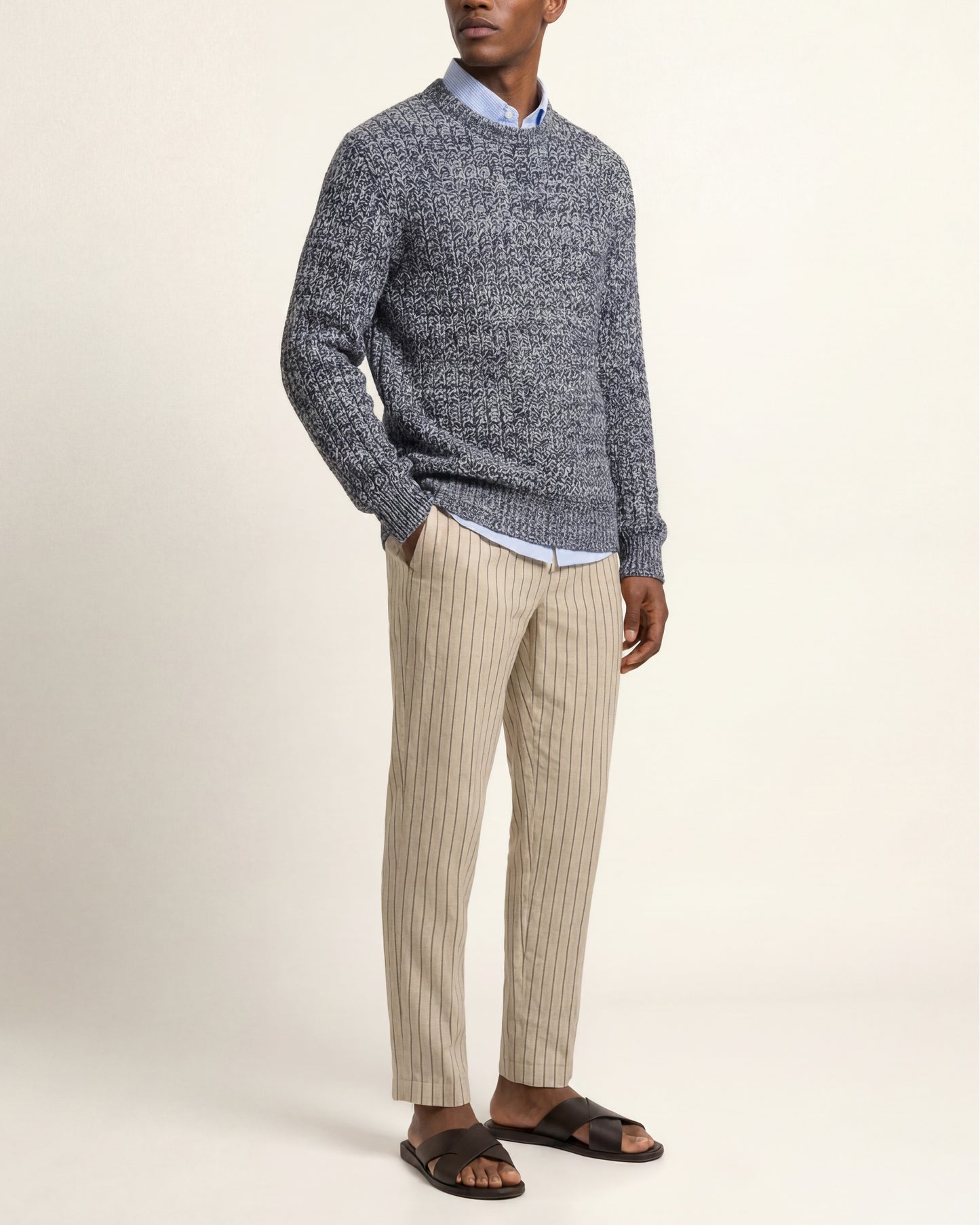 A man wearing a gray and navy marled knit sweater over a light blue collared shirt, paired with beige pinstriped trousers and dark brown slide sandals, standing against a plain cream background.
