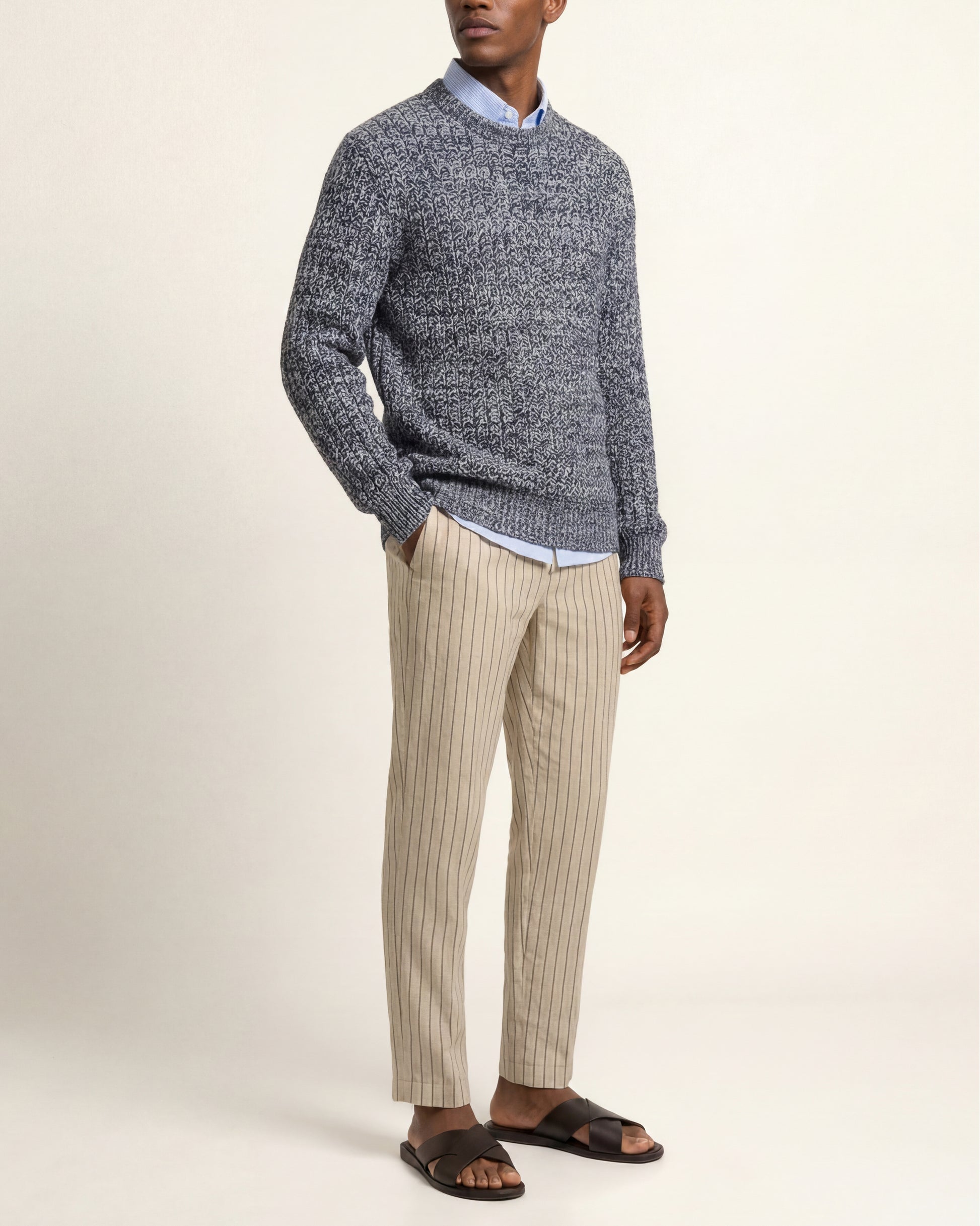 A man wearing a gray and navy marled knit sweater over a light blue collared shirt, paired with beige pinstriped trousers and dark brown slide sandals, standing against a plain cream background.