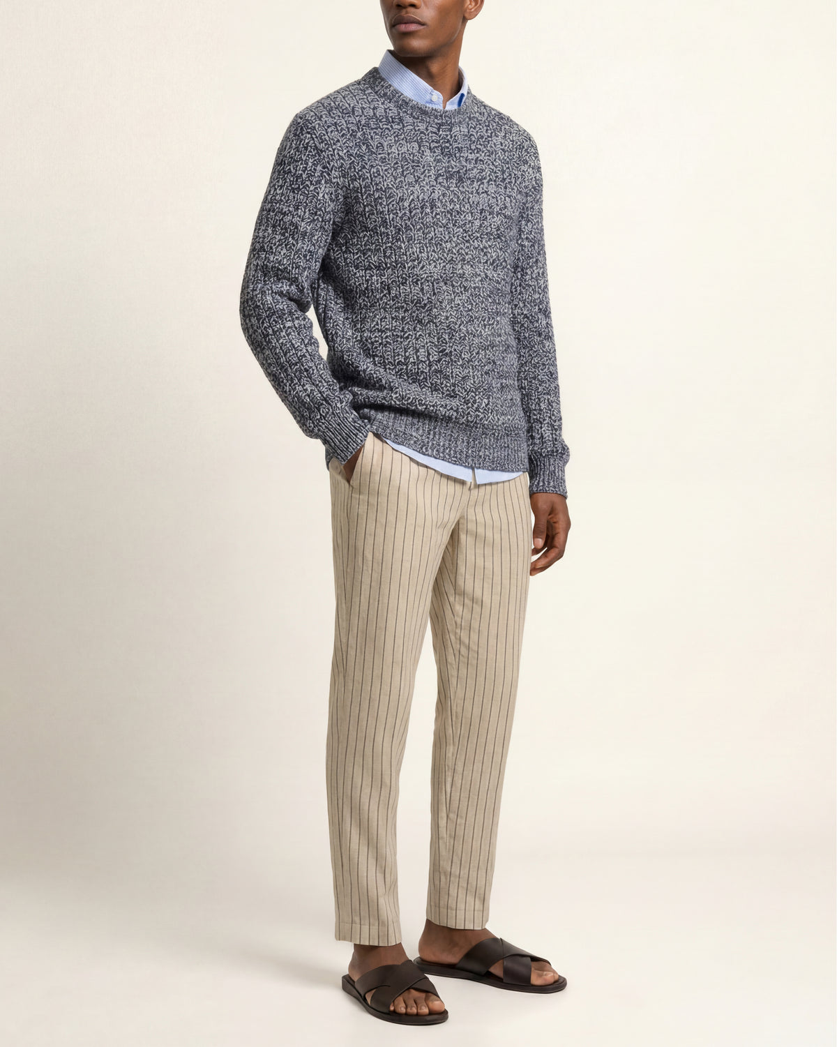 A man wearing a gray and navy marled knit sweater over a light blue collared shirt, paired with beige pinstriped trousers and dark brown slide sandals, standing against a plain cream background.