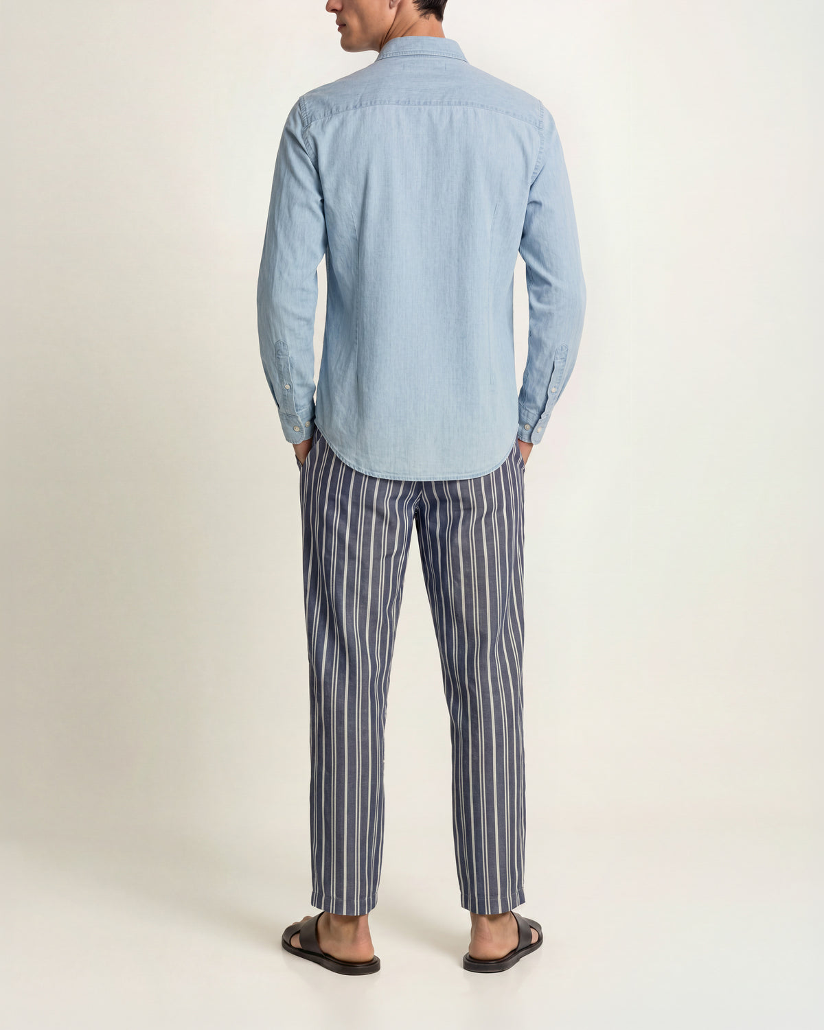 A man shown from the back wearing a light blue long-sleeve button-up shirt and navy-and-white vertically striped trousers paired with brown sandals standing against a plain studio background.