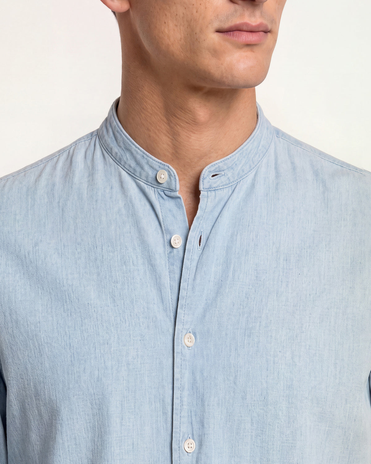 Close-up of a man wearing a light blue chambray button-down shirt with a mandarin collar and white buttons, set against a plain off-white background.