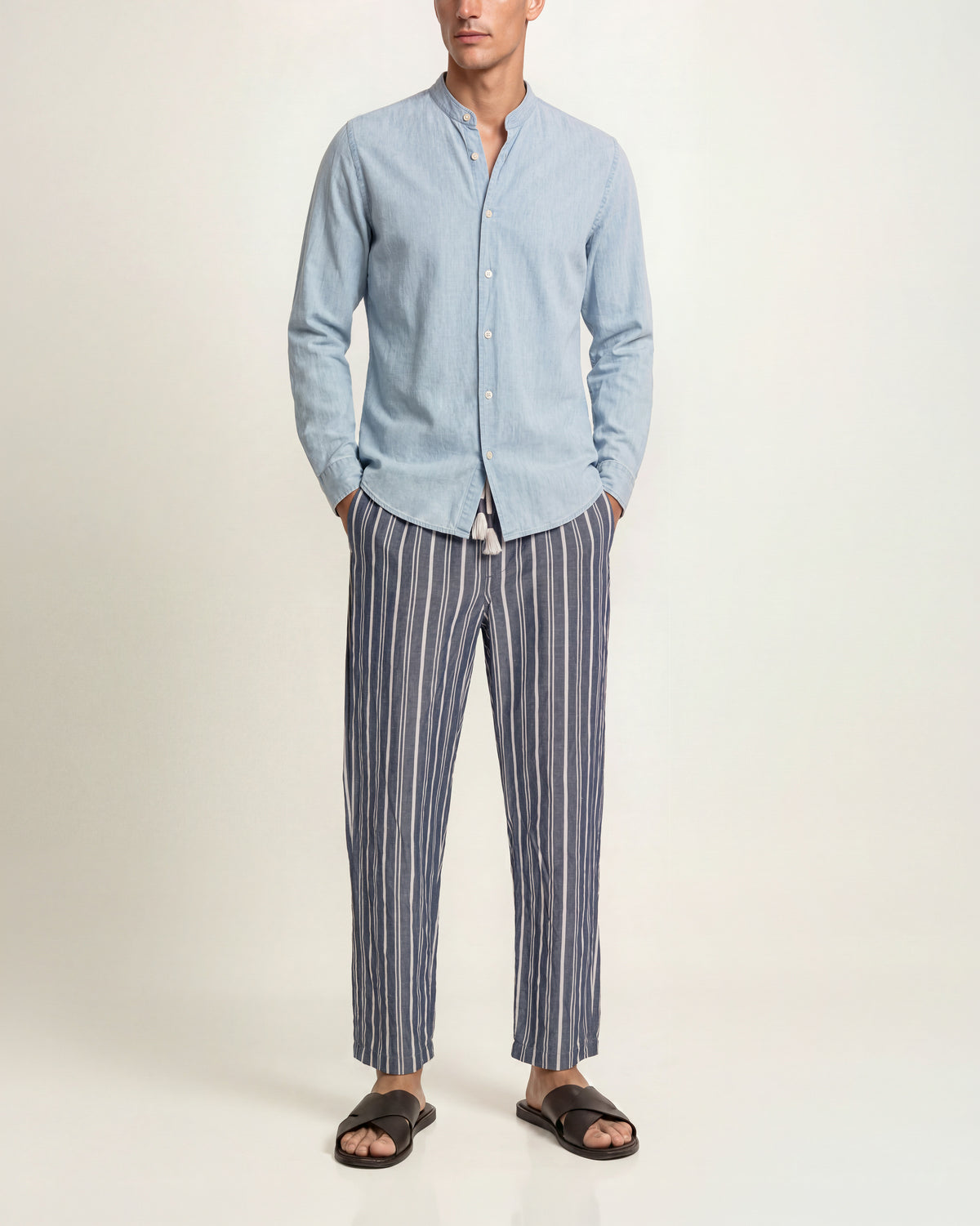 A man wearing a light blue long-sleeve button-up shirt with a stand collar and navy-and-white vertically striped trousers, paired with dark brown slide sandals, against a plain studio background.