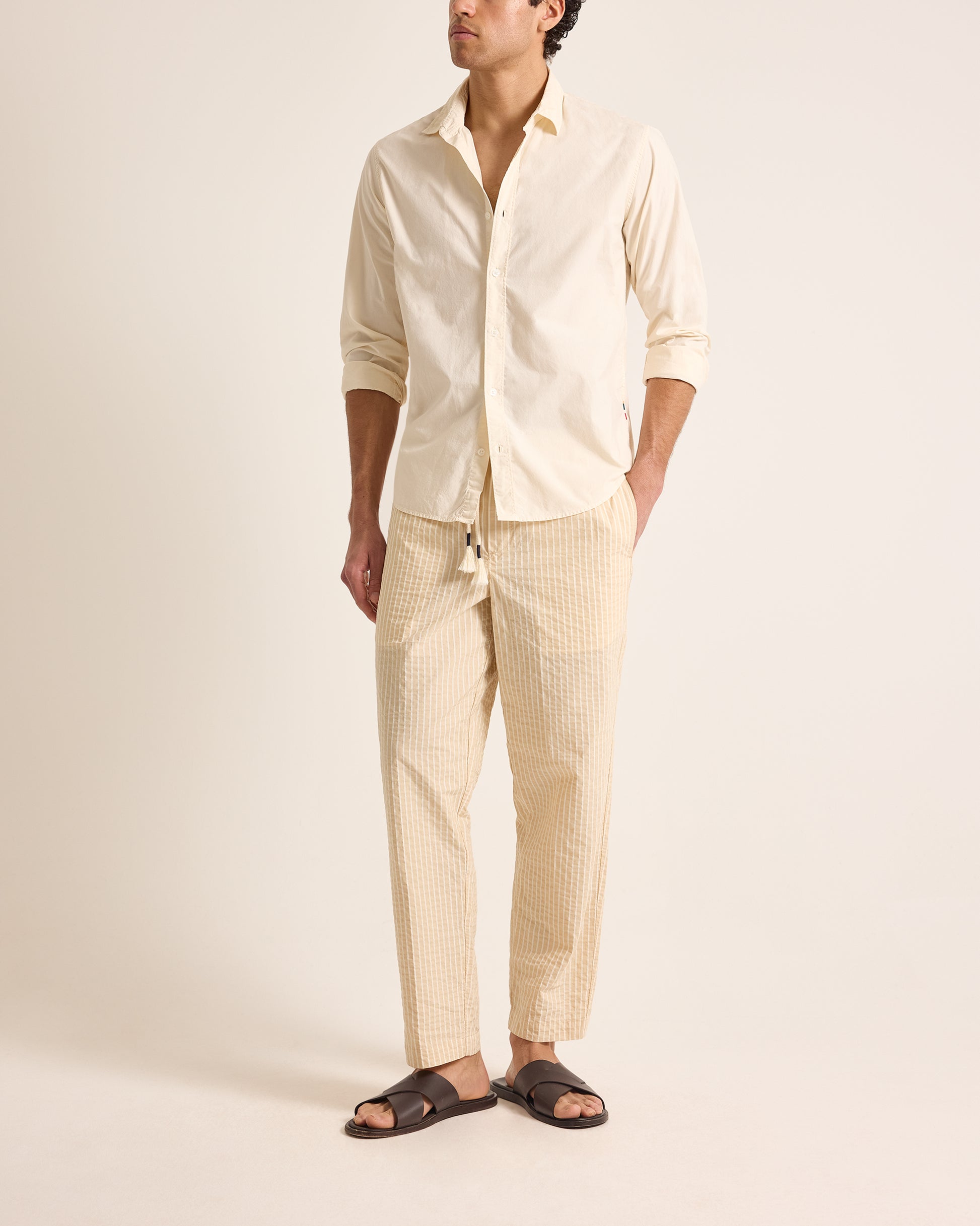 Man wearing a beige shirt and pants with sandals on a plain background