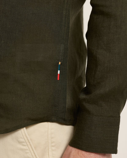 Close-up of a dark green shirt with a brand logo on the sleeve, worn over beige pants.