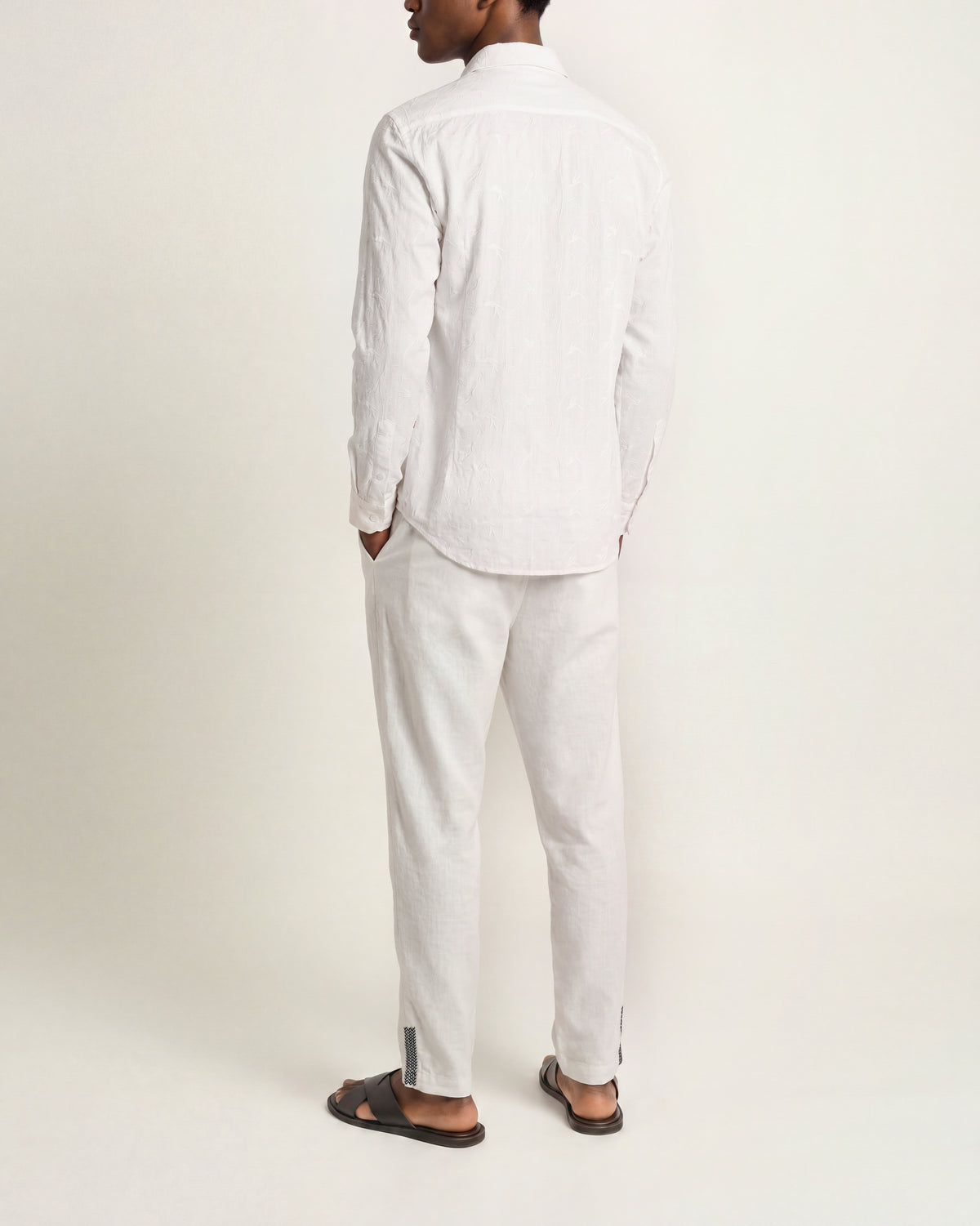 Back view of a man wearing white long-sleeved shirt with embroidery and white drawcord waist trousers and black sandals on a plain background