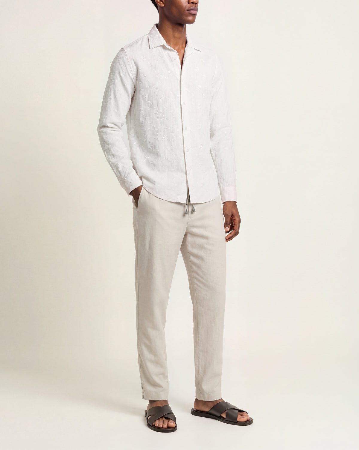 A man modeling a white long-sleeve linen shirt and light beige linen pants with dark brown slide sandals, standing against a plain off-white studio background