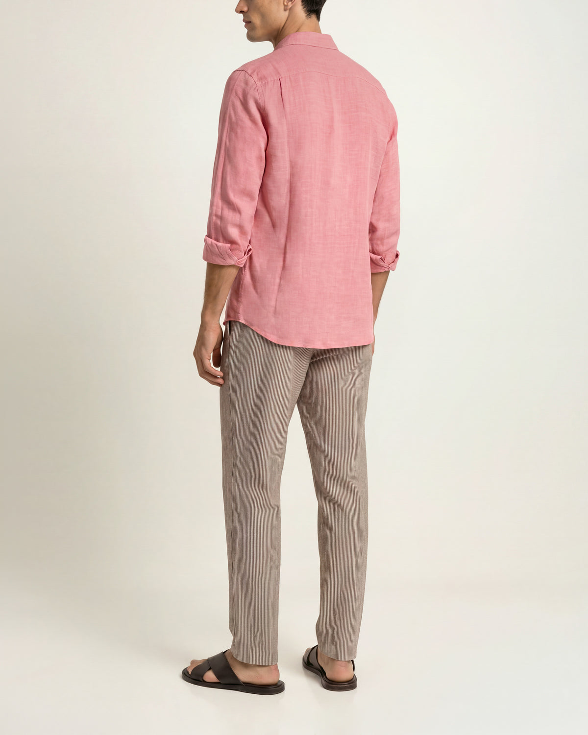 Back view of a man wearing a light red long-sleeved shirt paired with white-and-black patterned drawcord-waist trousers and brown sandals on a plain background