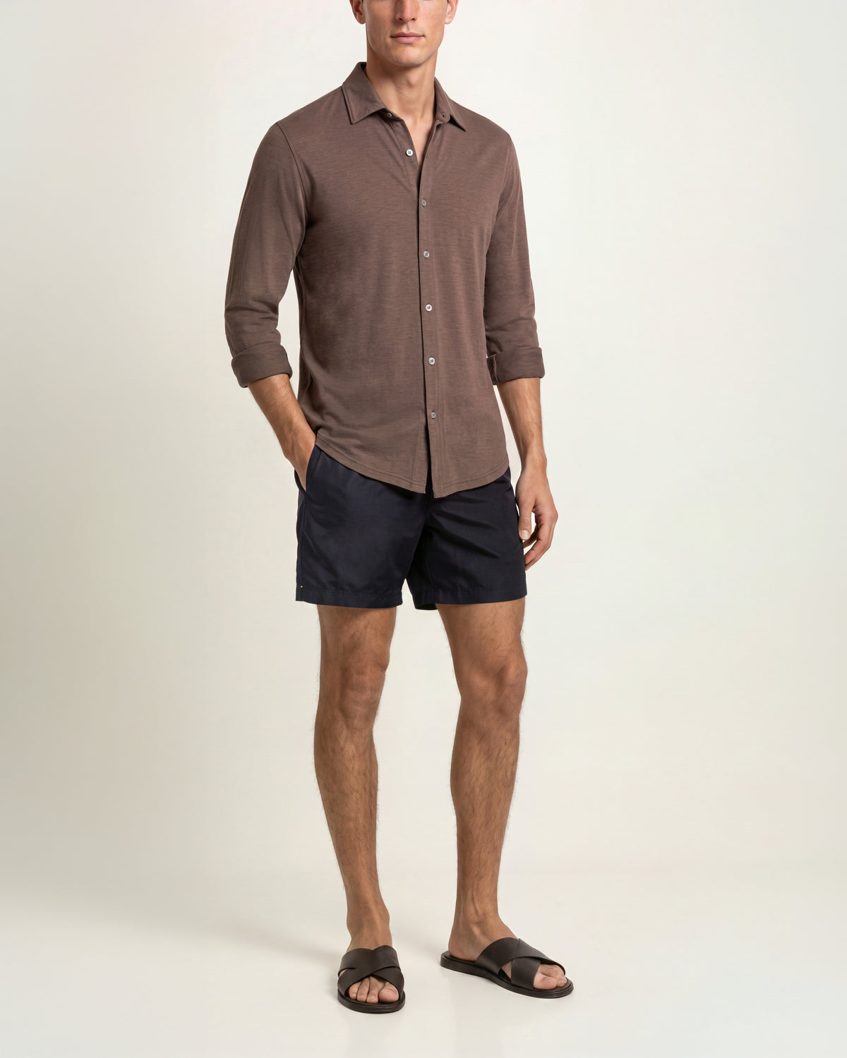 Man wearing brown shirt with black shorts and brown sandals on a plain background