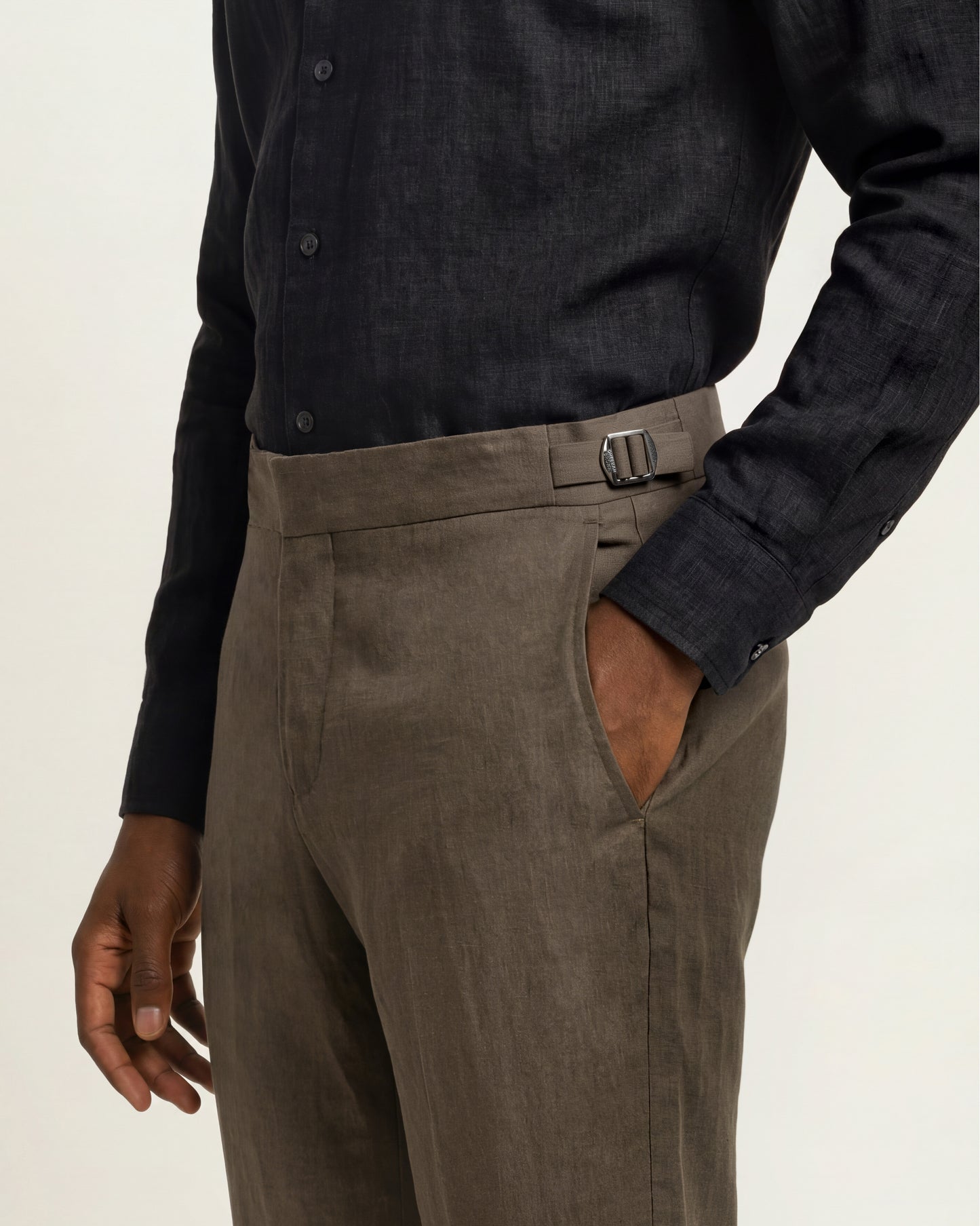 Close-up of a person wearing tailored trousers in a muted taupe-brown tone with a matching belt, paired with a dark charcoal button-up shirt, against a plain light background.
