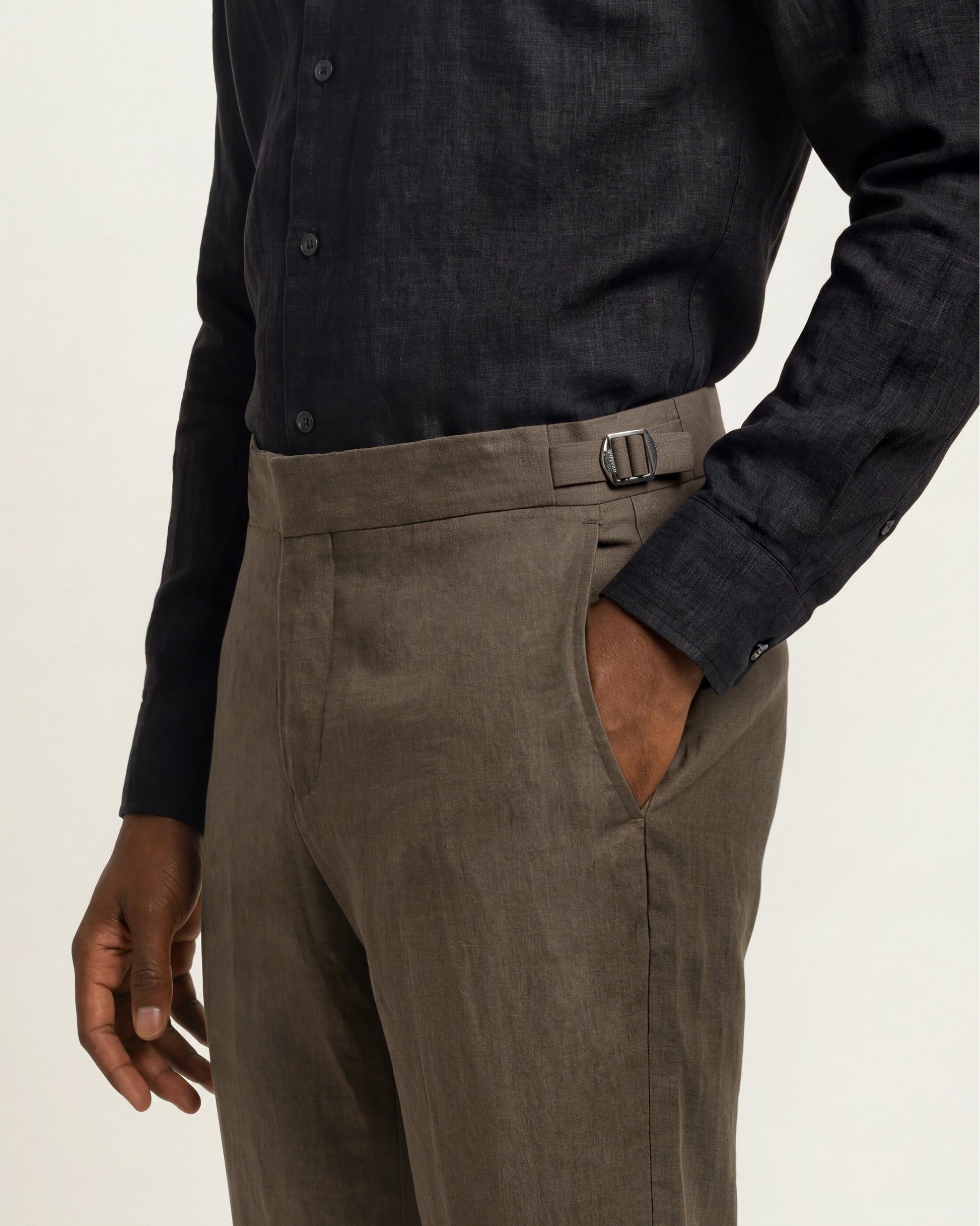Close-up of a person wearing tailored trousers in a muted taupe-brown tone with a matching belt, paired with a dark charcoal button-up shirt, against a plain light background.