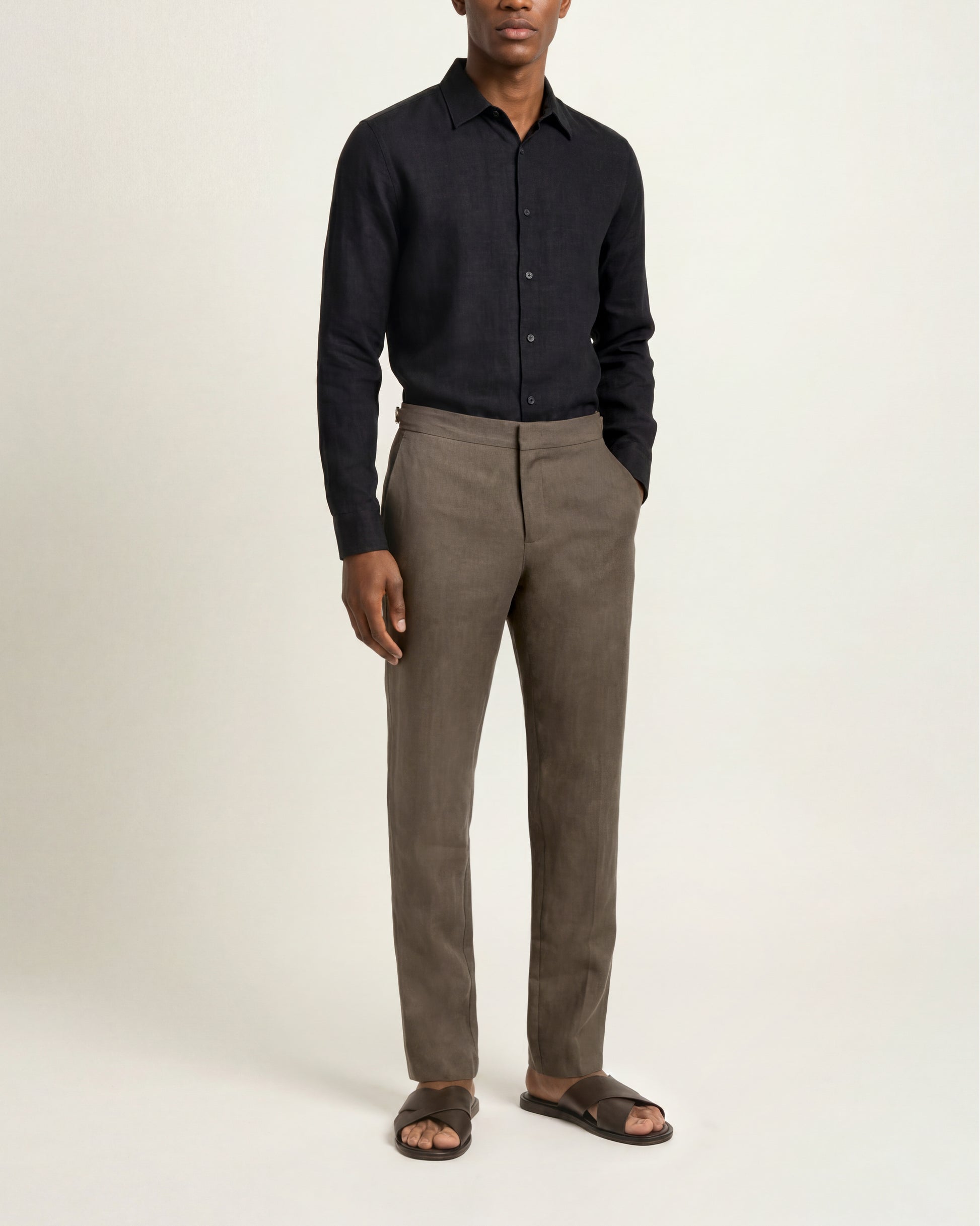 A man modeling a dark navy long-sleeve button-down shirt and taupe-brown straight-leg dress pants, paired with dark brown slide sandals, against a plain off-white studio background.