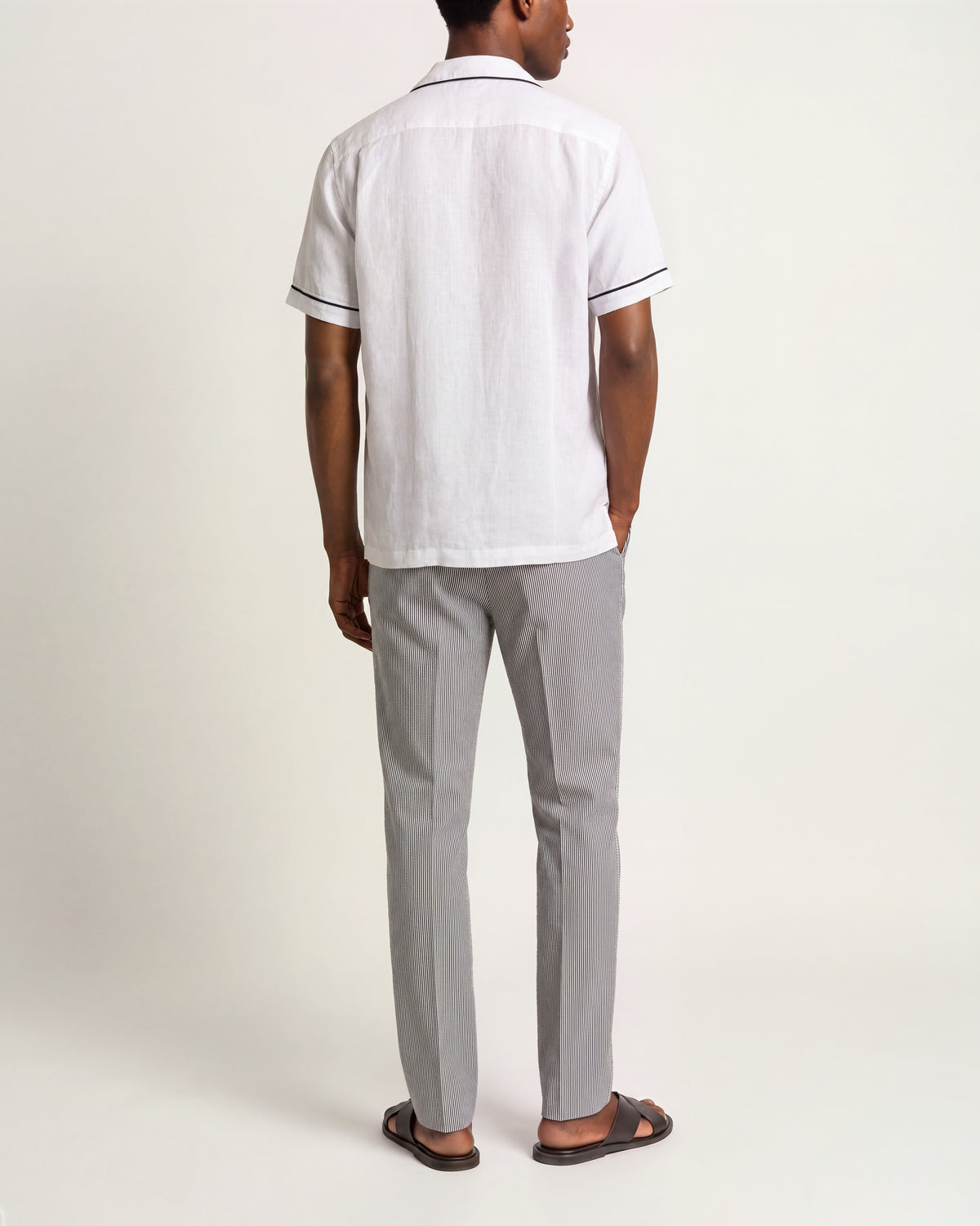 A man viewed from the back wearing a white short-sleeve linen shirt with thin dark piping on the collar and sleeve cuffs, paired with light gray trousers and dark brown sandals, against a plain off-white studio background.