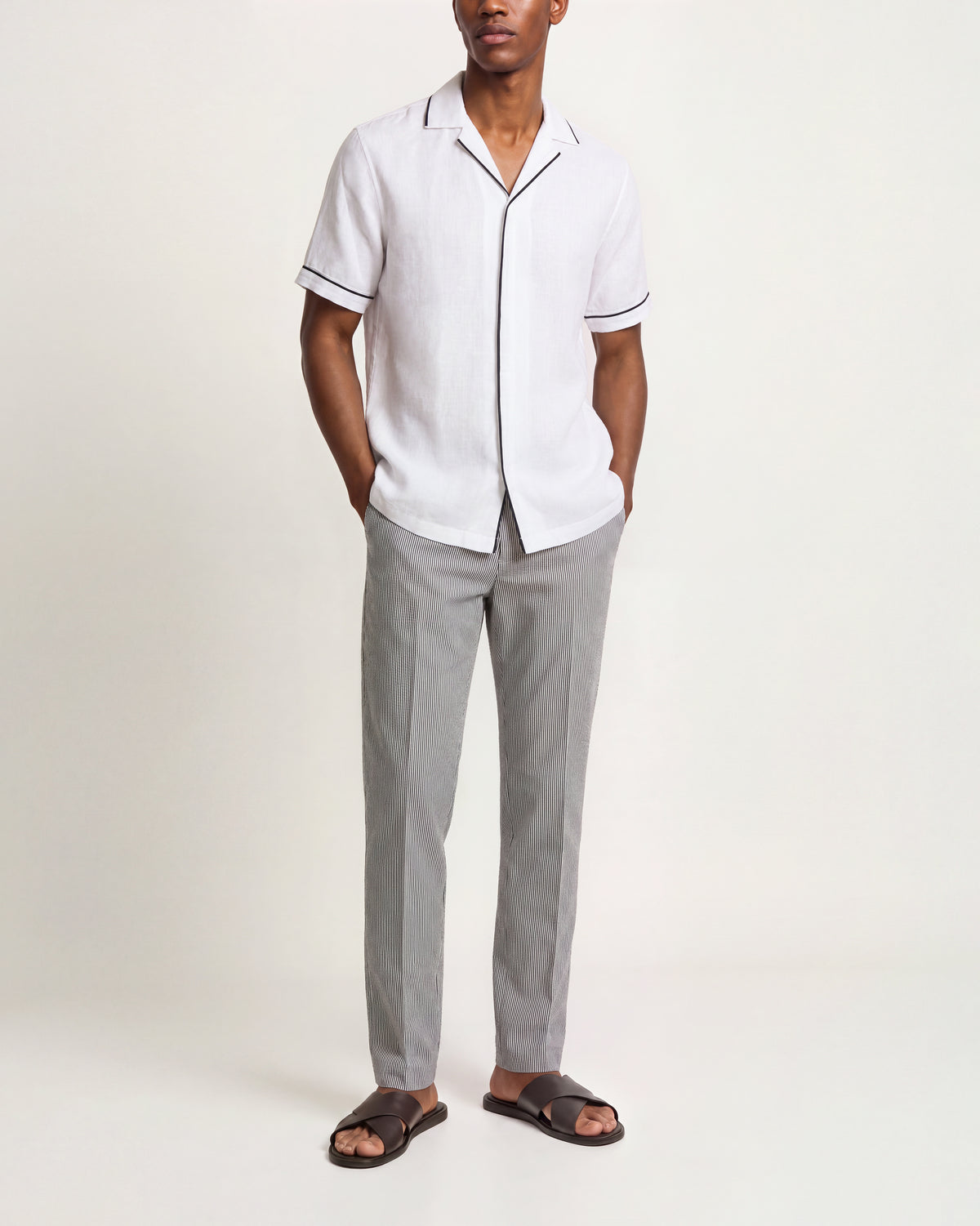 A man modeling a white short-sleeve button-up shirt with dark piping trim on the collar and sleeve cuffs, paired with light gray trousers and dark brown slide sandals, against a plain off-white studio background.