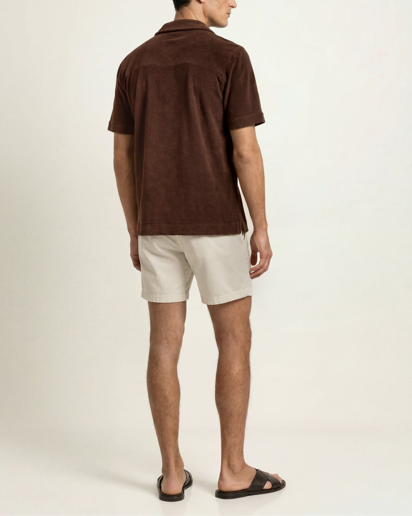 A man shown from the back wearing a brown short-sleeve collared shirt, off-white shorts, and black slide sandals against a plain light brown studio background.